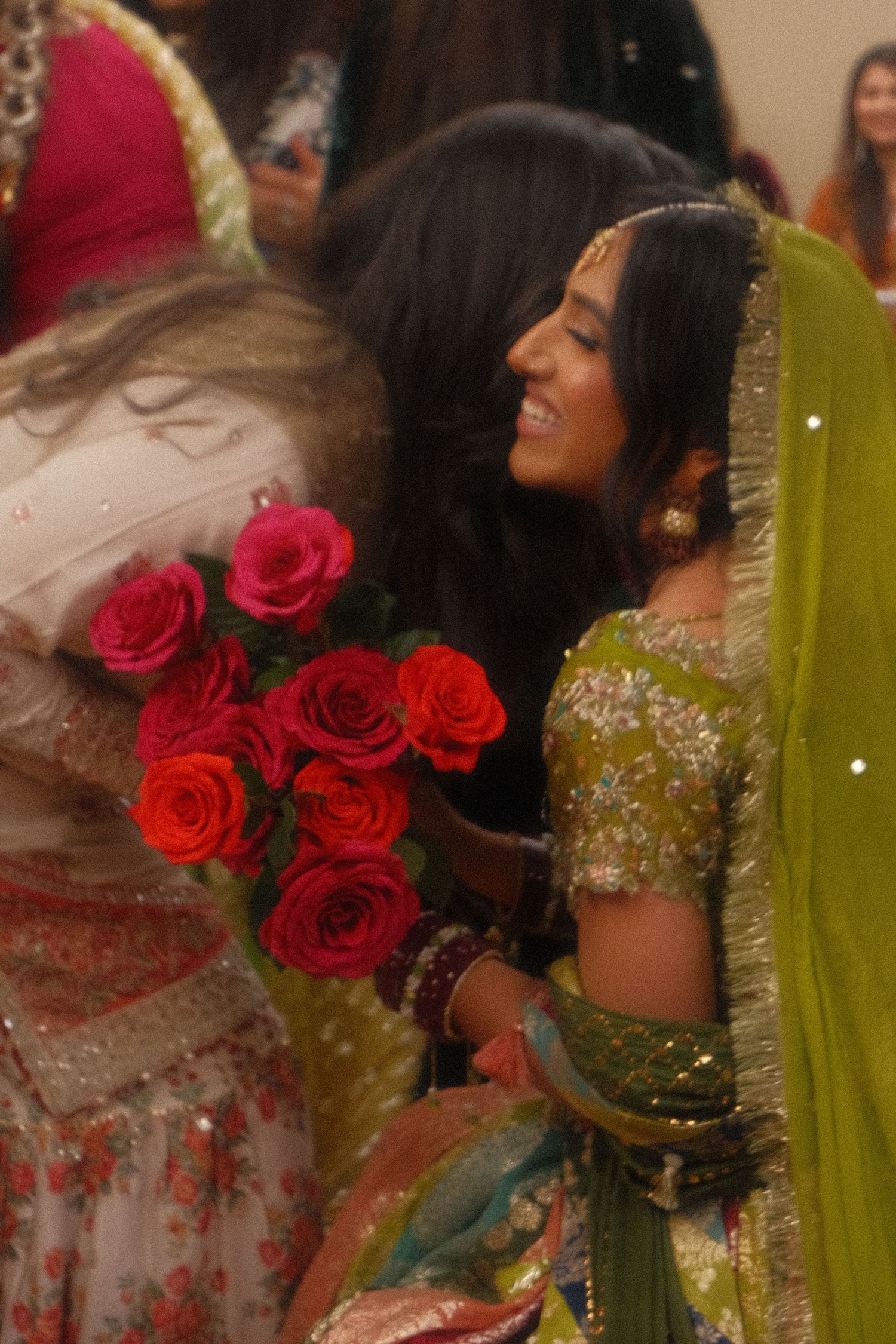 Smiling bride in green embroidered lehenga with dupatta receiving red roses bouquet at South Asian wedding celebration.
