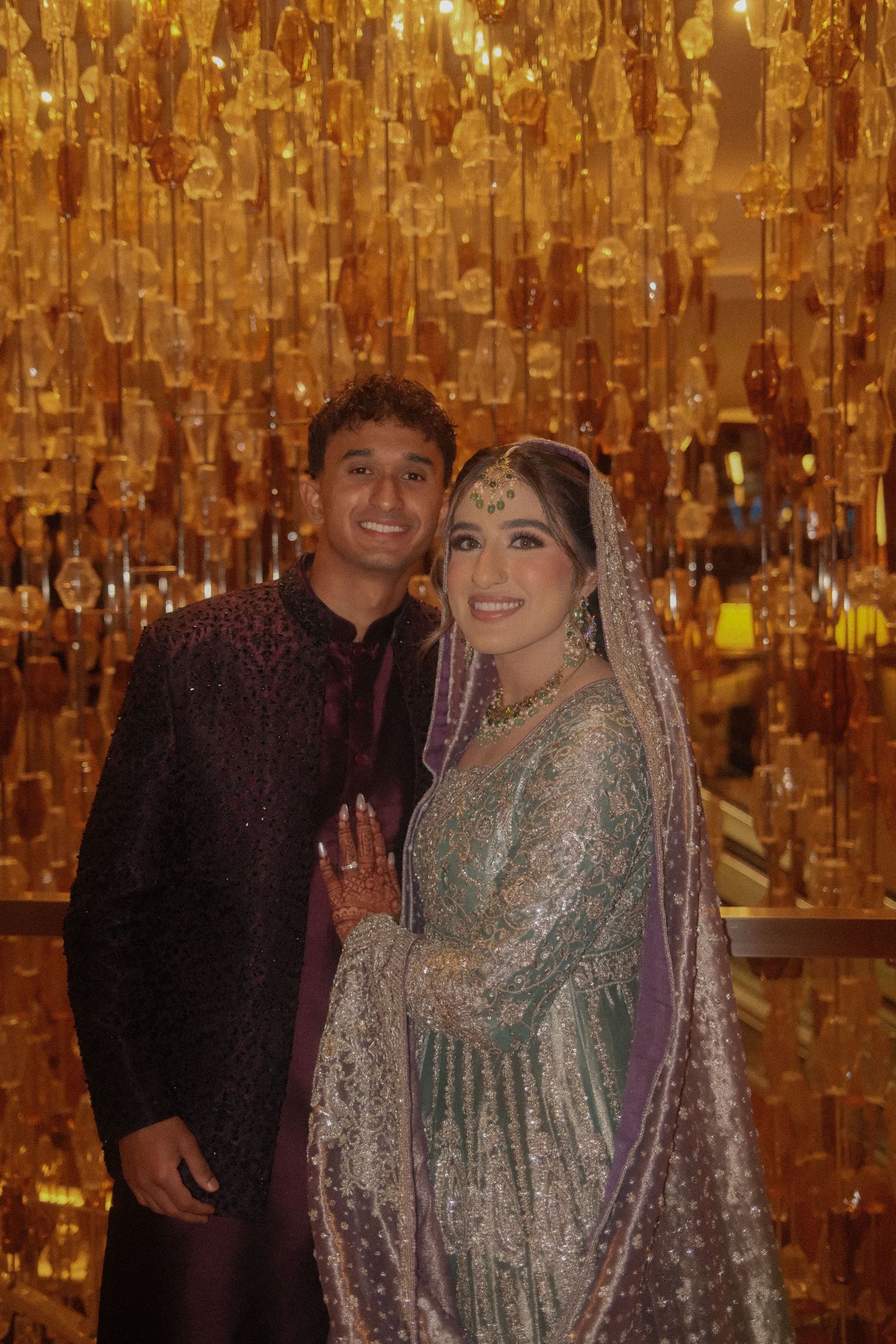 South Asian bride and groom posing under golden chandelier, bride in teal embroidered lehenga with purple dupatta.