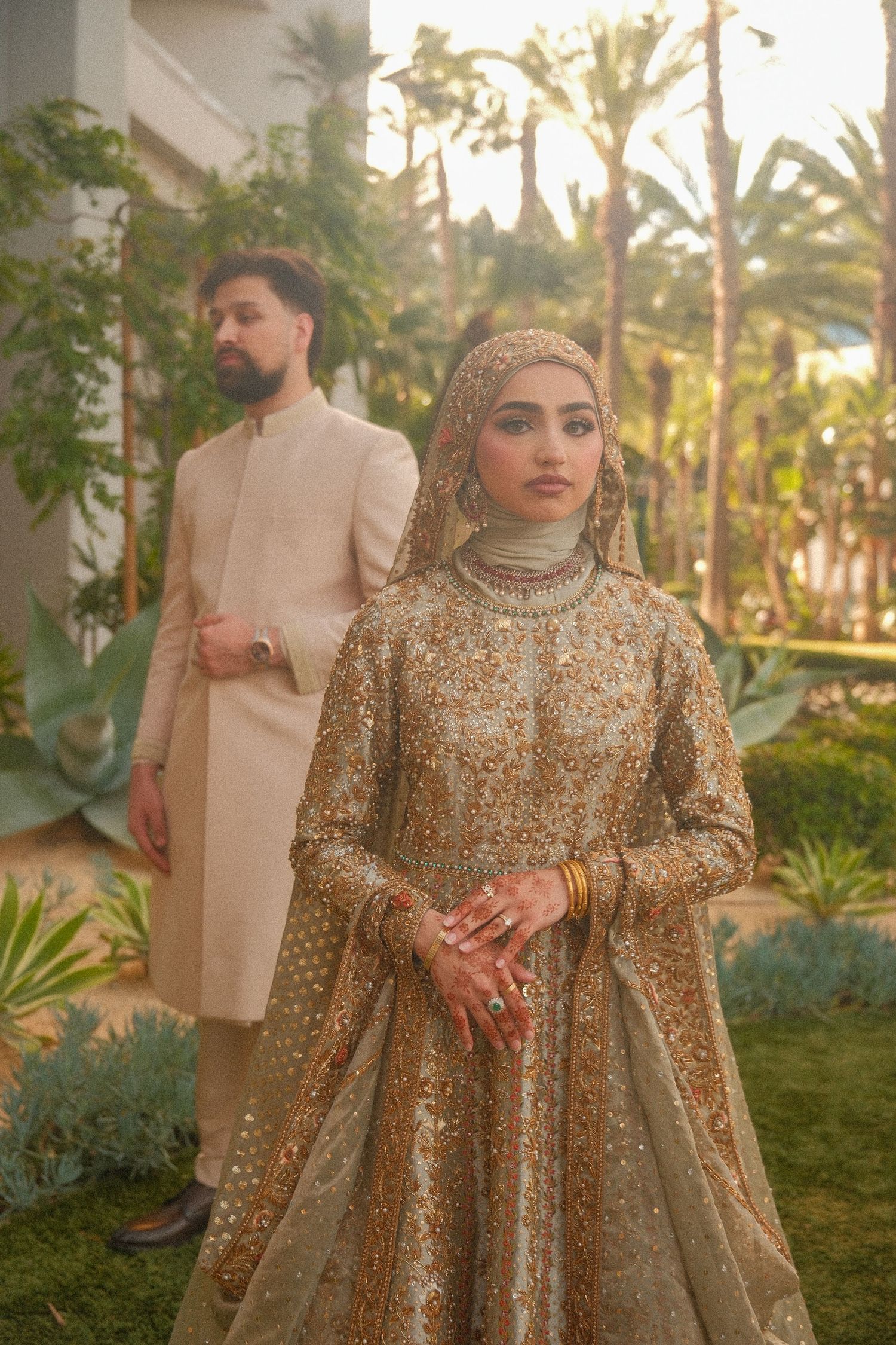 Pakistani bride in gold embroidered lehenga and groom in cream sherwani at outdoor garden wedding.