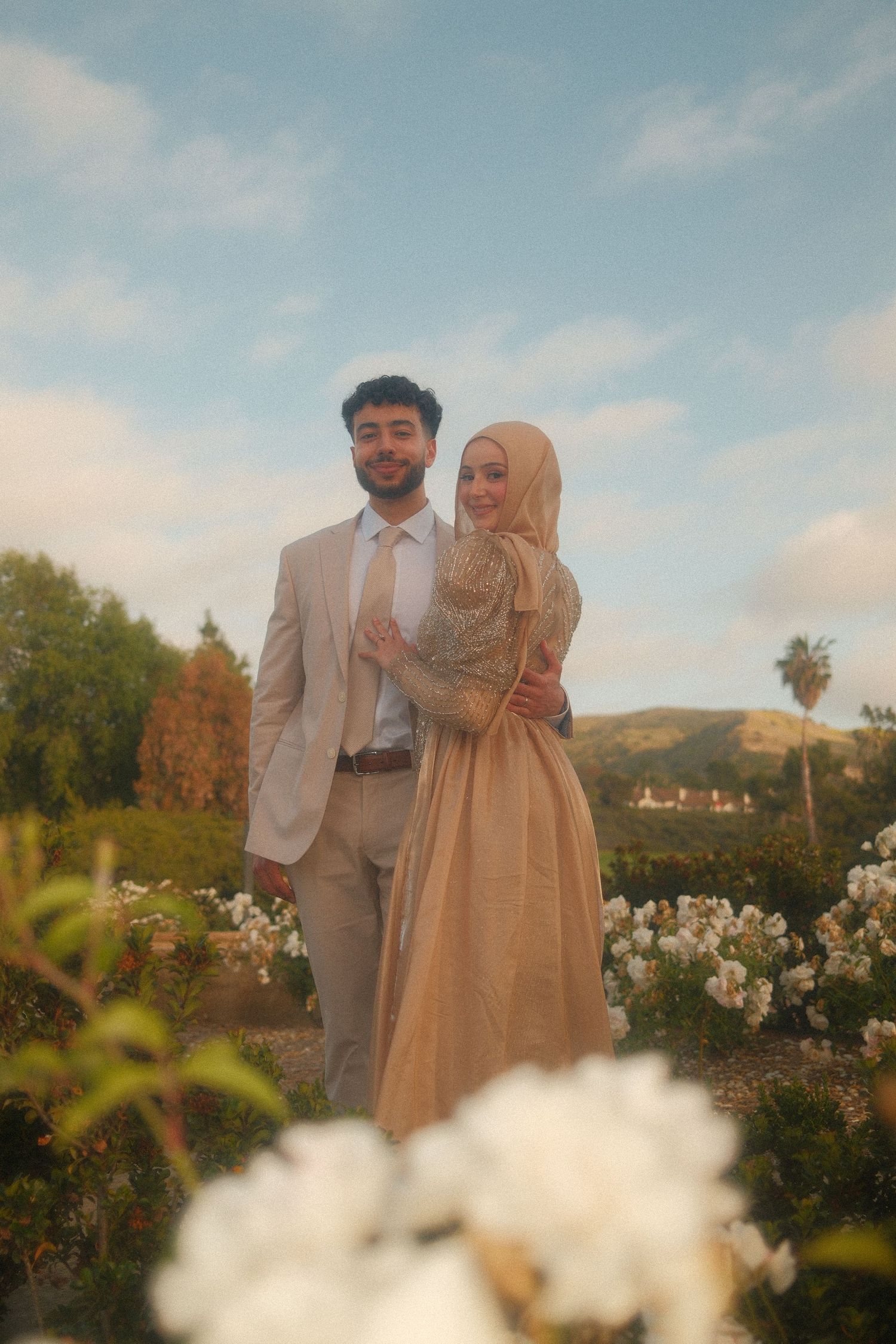 A couple in matching gold outfits pose in a garden with white flowers and hills in the background.