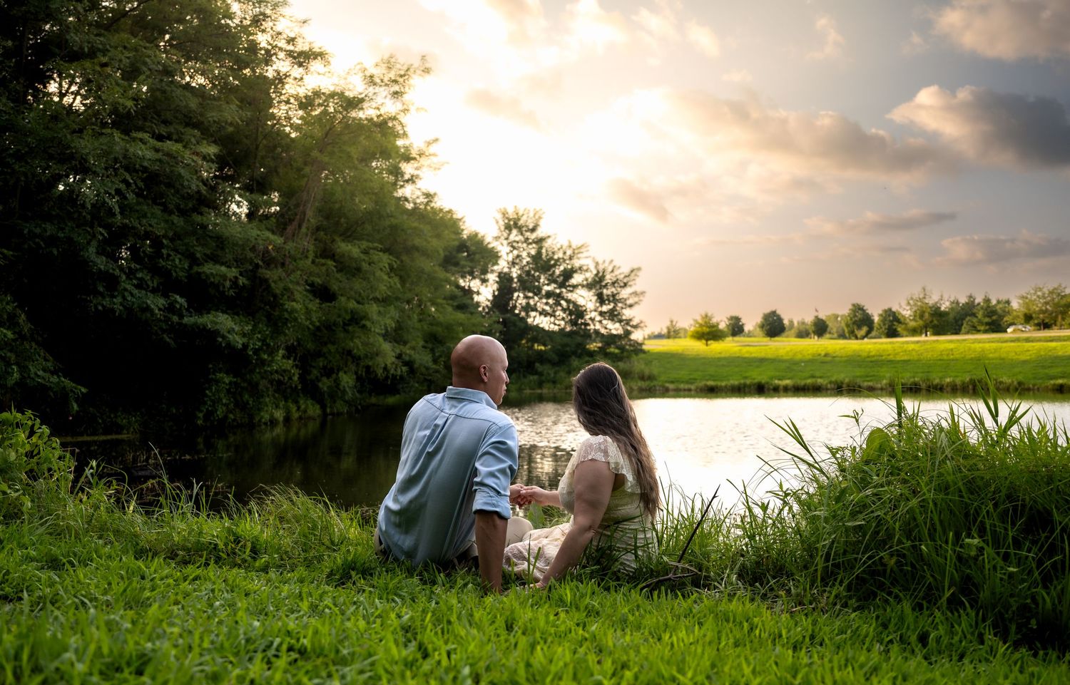 lifestyle portrait of a Couple sitting together on lush green grass by a tranquil pond at sunset, surrounded by trees.
