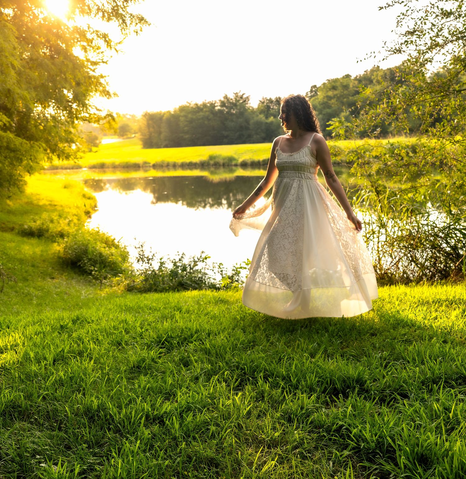 lifestyle portrait of woman who stands by a sunlit pond surrounded by lush green grass and trees at golden hour.