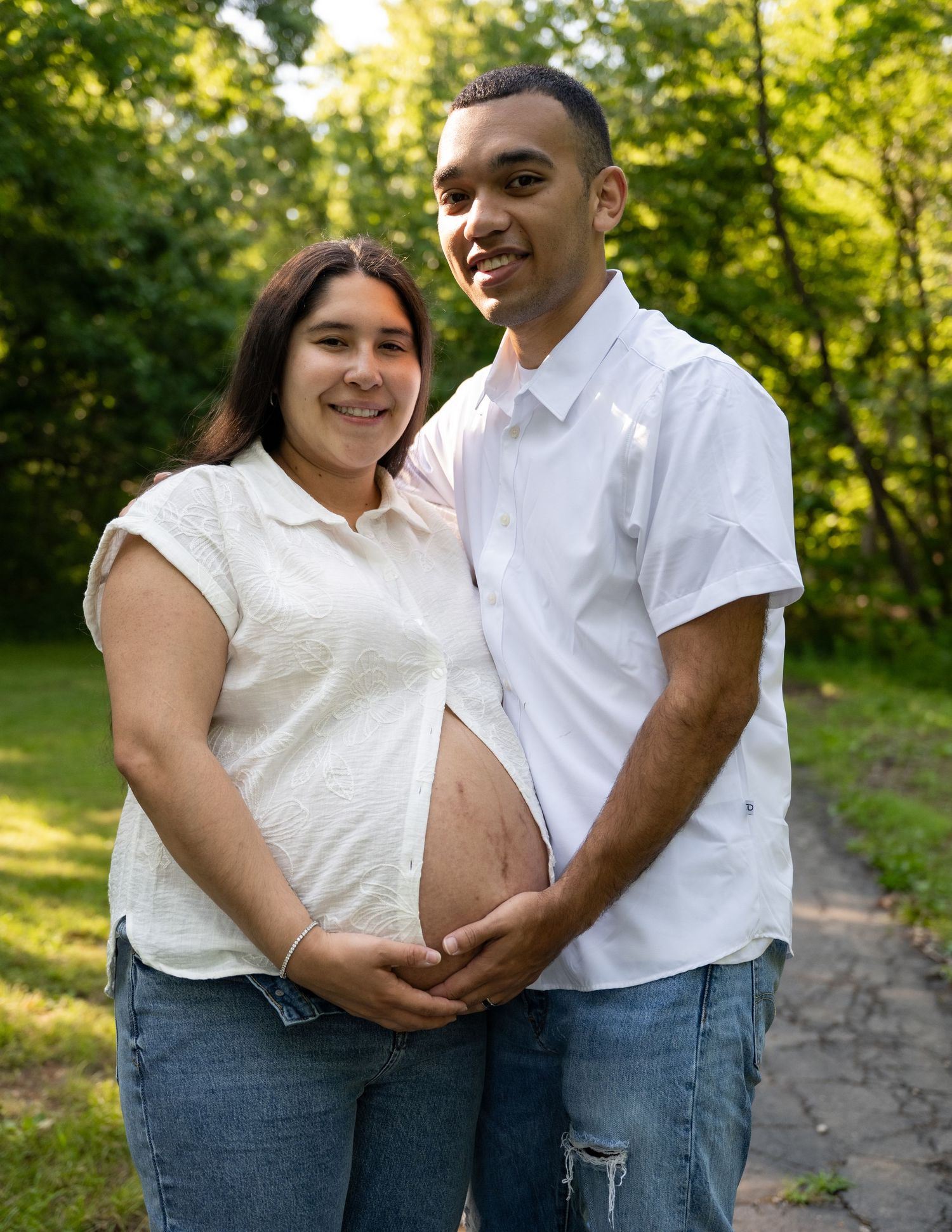 portrait of a Smiling expectant couple posing outdoors on a tree-lined path, cradling pregnant belly.