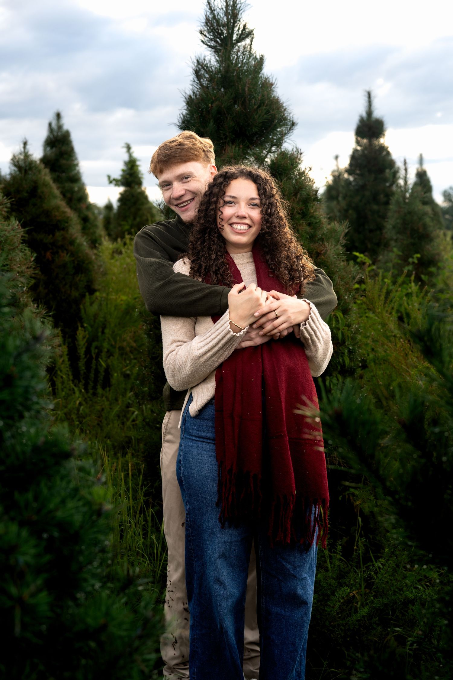 A happy couple embraces among green Christmas trees at a tree farm, the man hugging the woman from behind as she smiles.