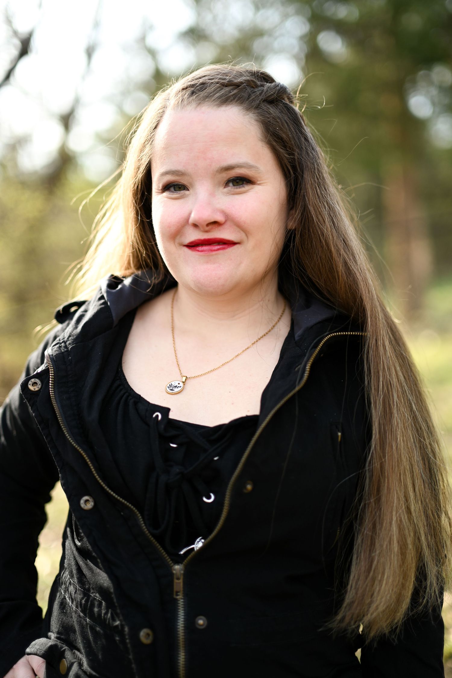 Smiling young woman with long hair wearing black jacket outdoors in natural setting.
