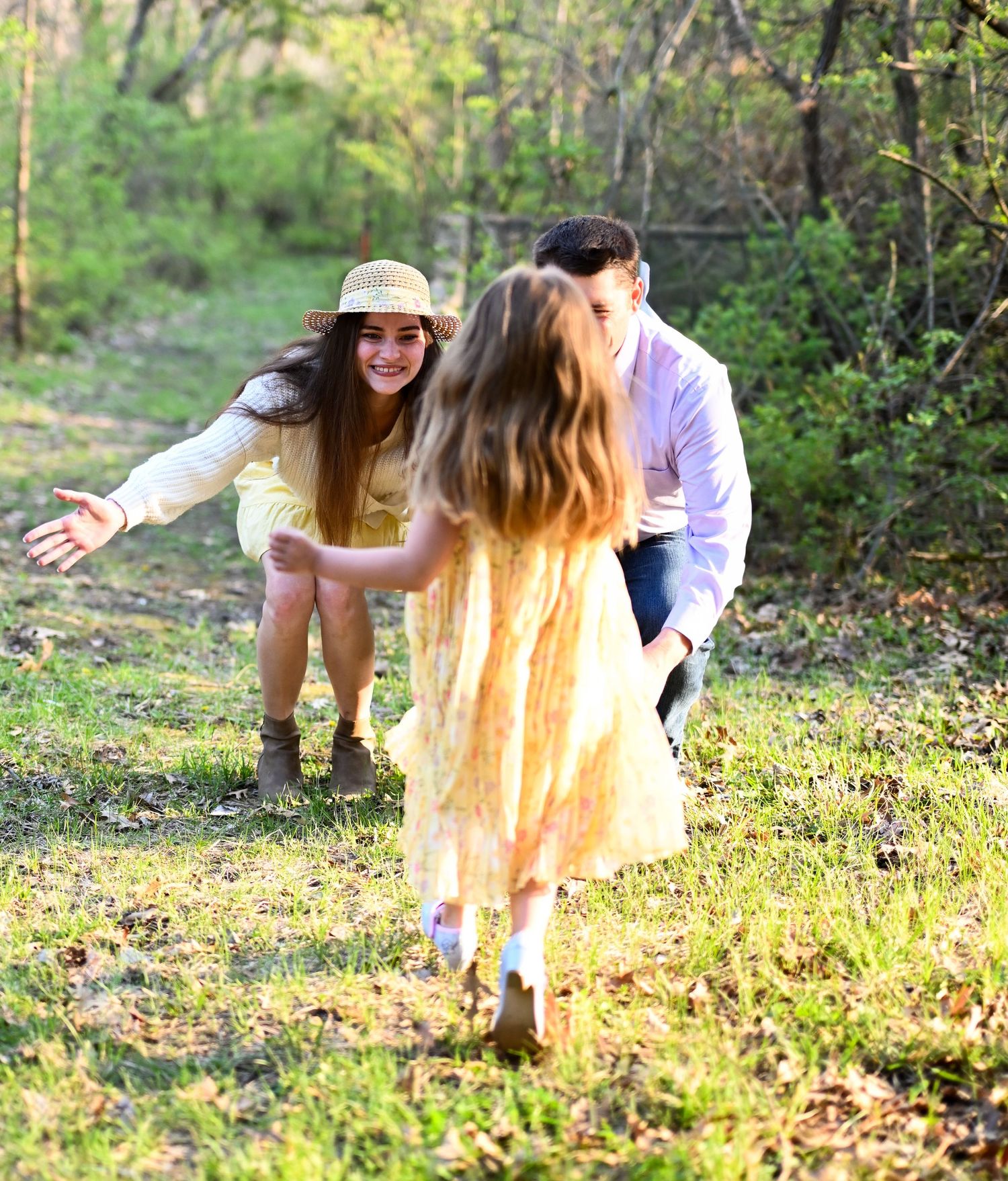 A young girl runs toward a smiling woman in a straw hat and man in a sunny outdoor field.