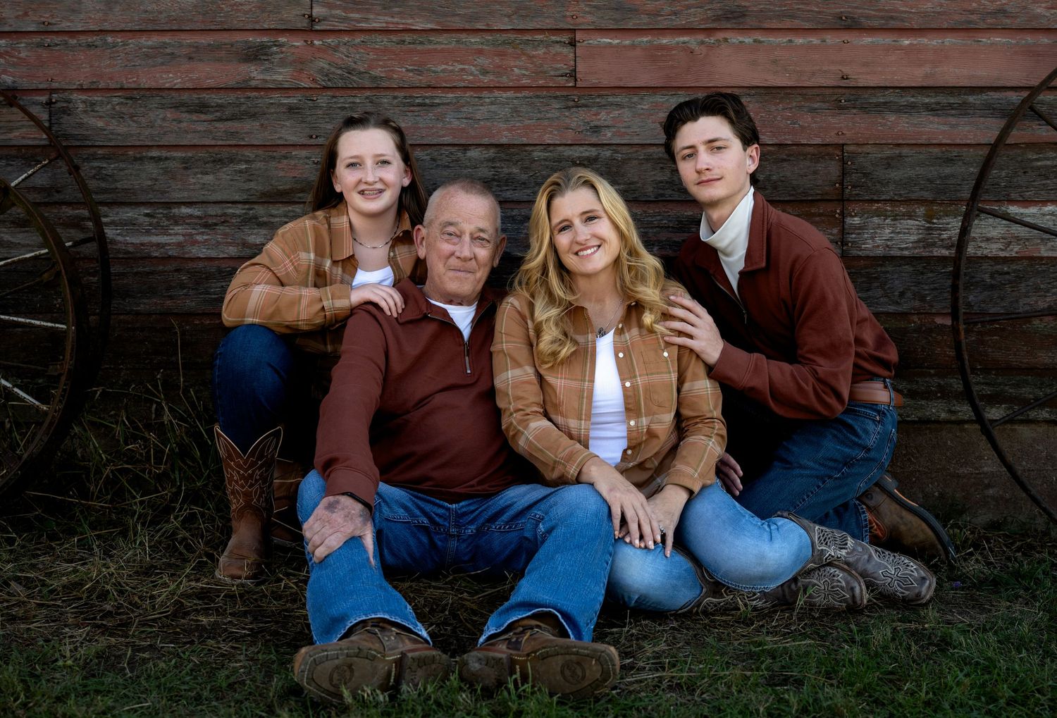 Four family members in denim and rust-toned shirts pose together outdoors on grass in front of a rustic wooden barn wall and wagon wheel.
