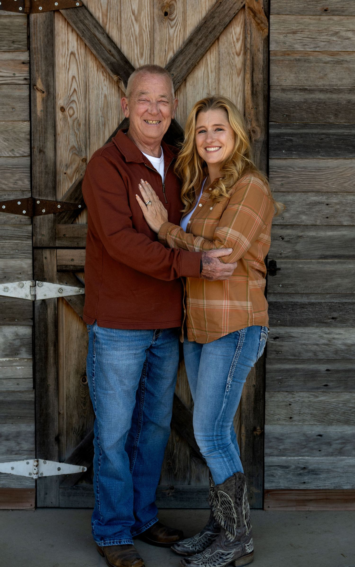 Smiling older man in a rust jacket embraces a blonde woman in a plaid shirt and cowboy boots in front of a rustic wooden barn door.