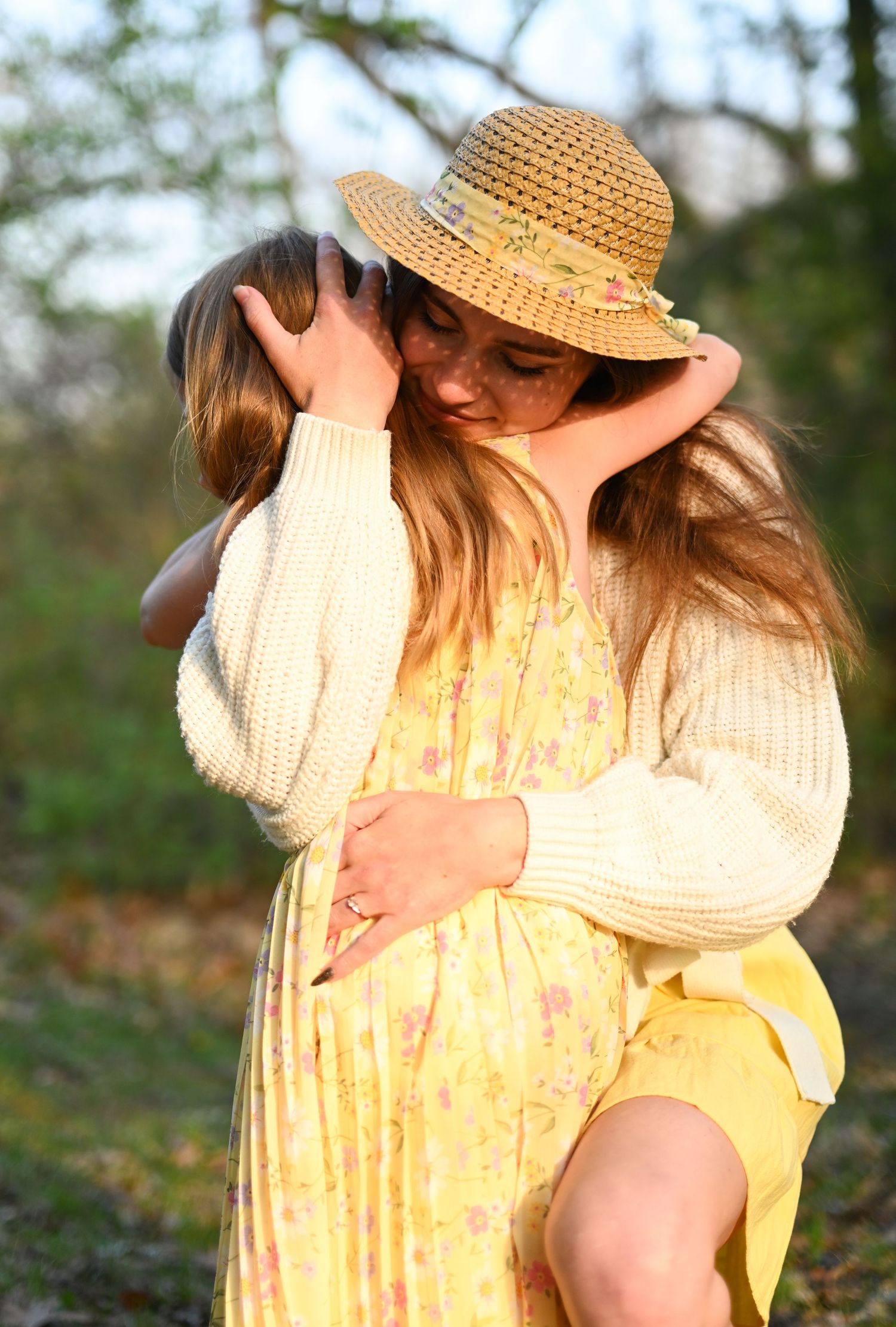Mother in yellow floral dress and straw hat embracing young daughter in nature.