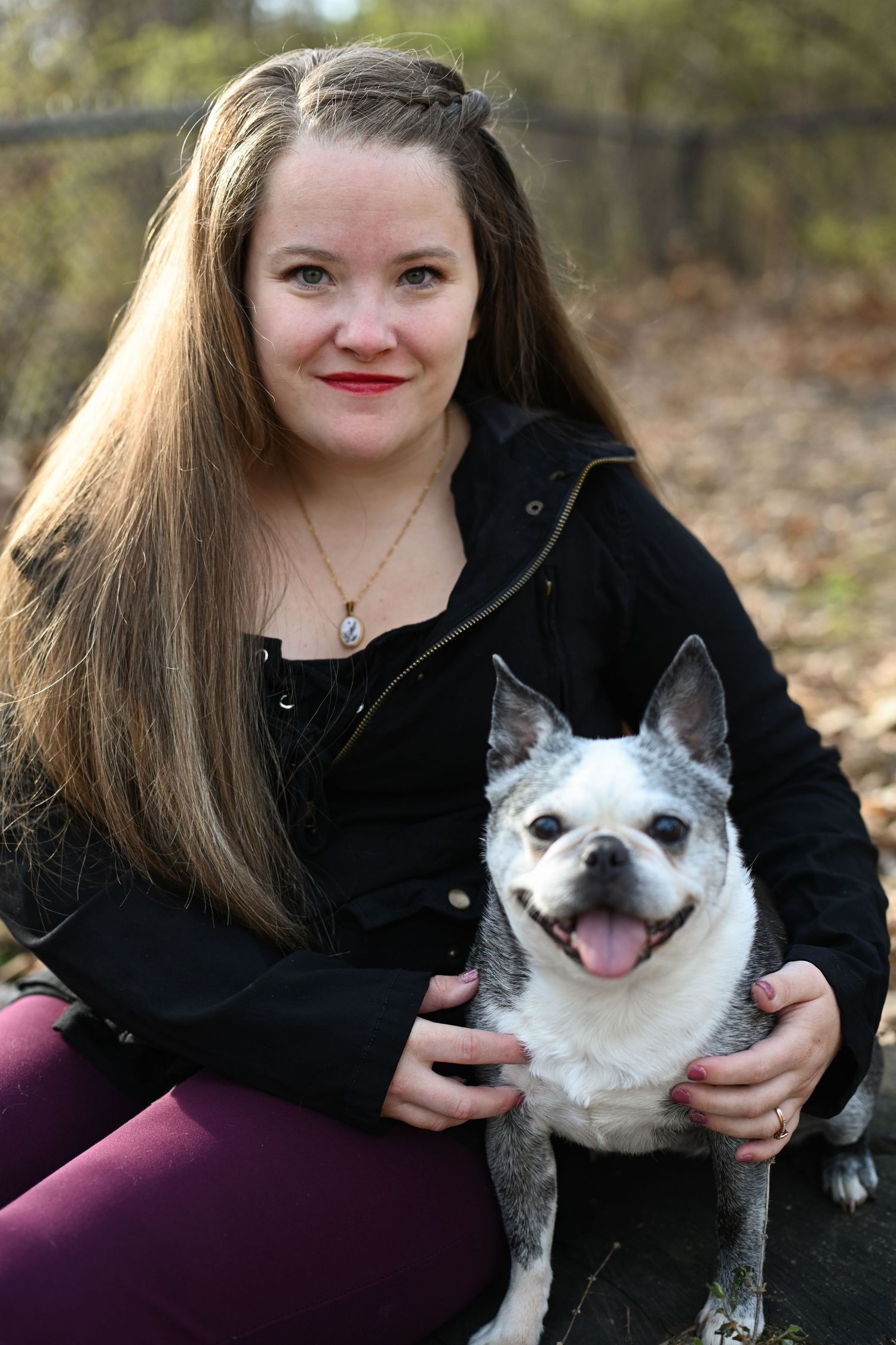 Smiling young woman with long hair posing with Boston Terrier dog outdoors.