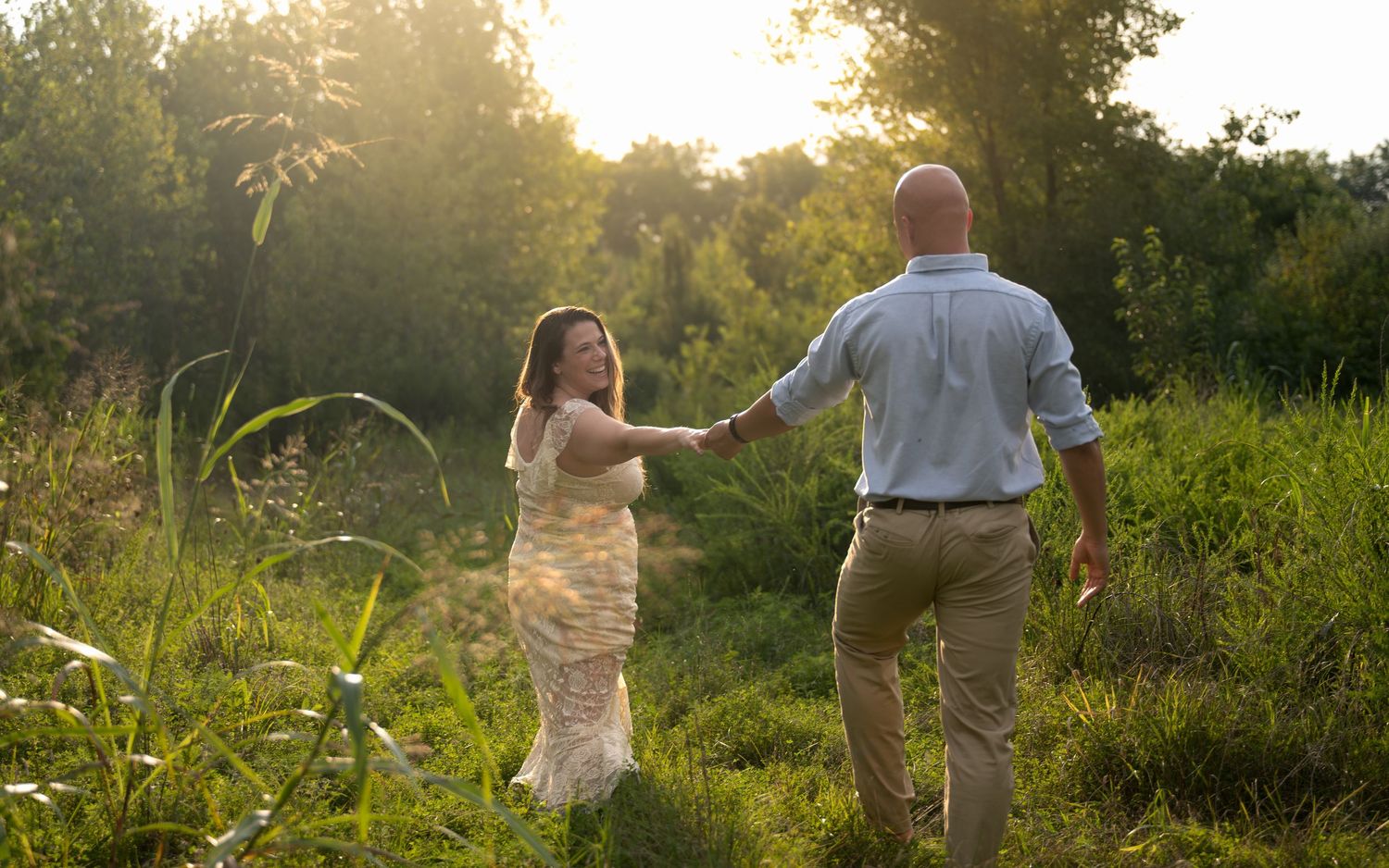 lifestyle portrait of a woman in a floral dress holds hands with a man in a sunlit green meadow at golden hour.