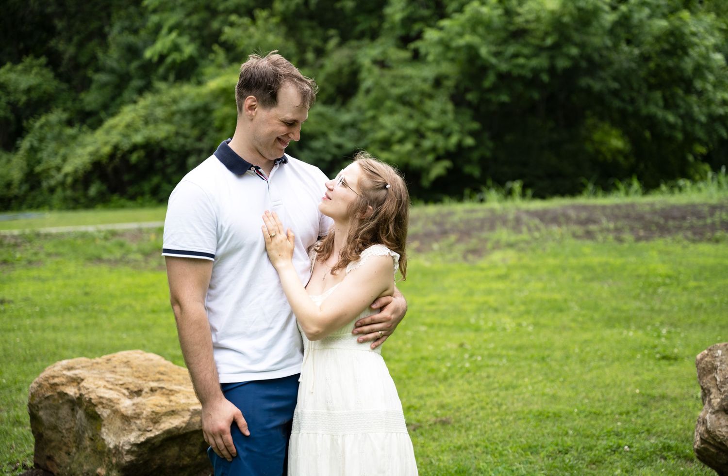 portrait of a Young couple in white outfits share a tender moment on a green lawn with large rocks.