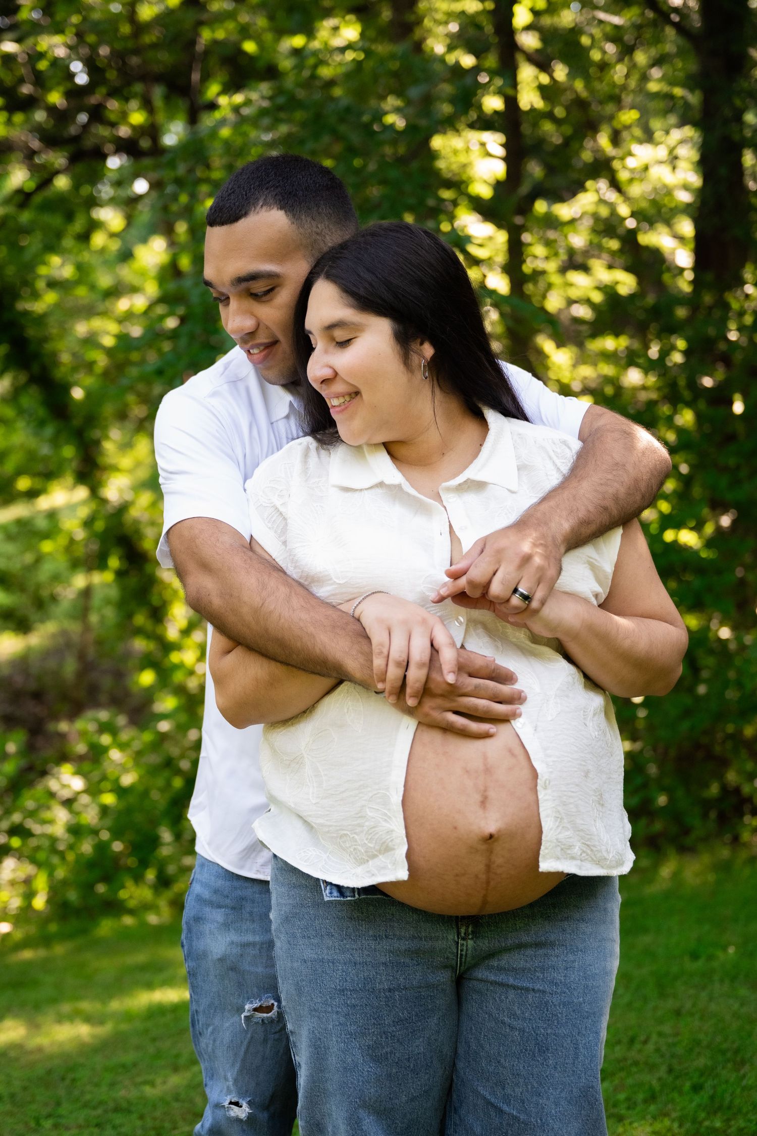 portrait of a Loving couple embracing outdoors, man hugging pregnant woman from behind in a green park.