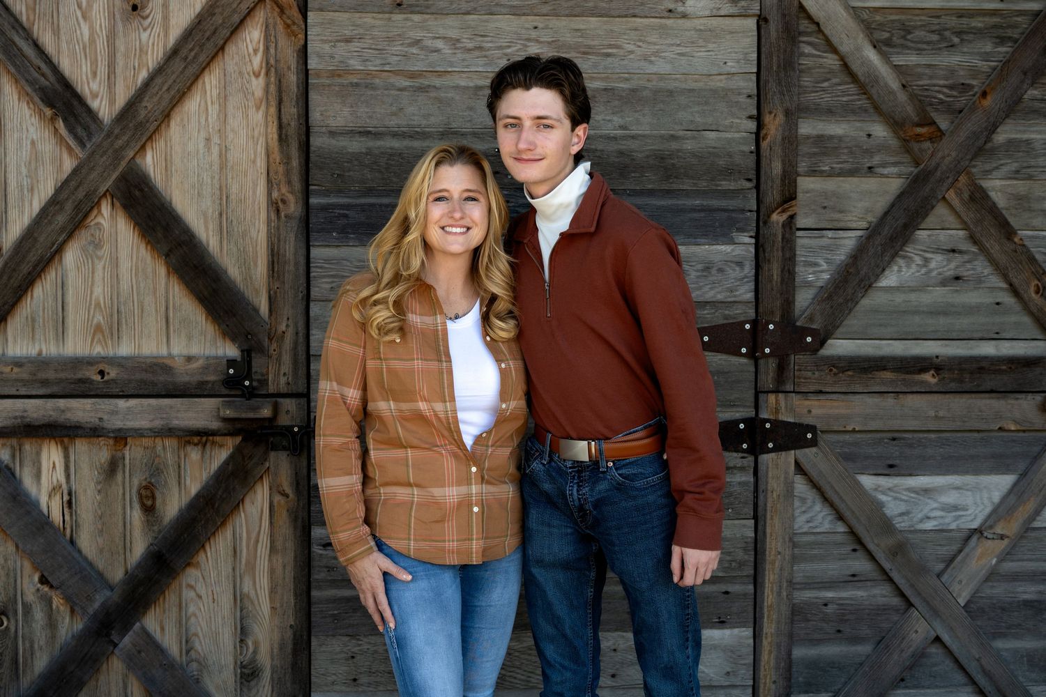Smiling young woman in a plaid shirt and young man in a rust-red sweater posing together in front of a rustic wooden barn door.