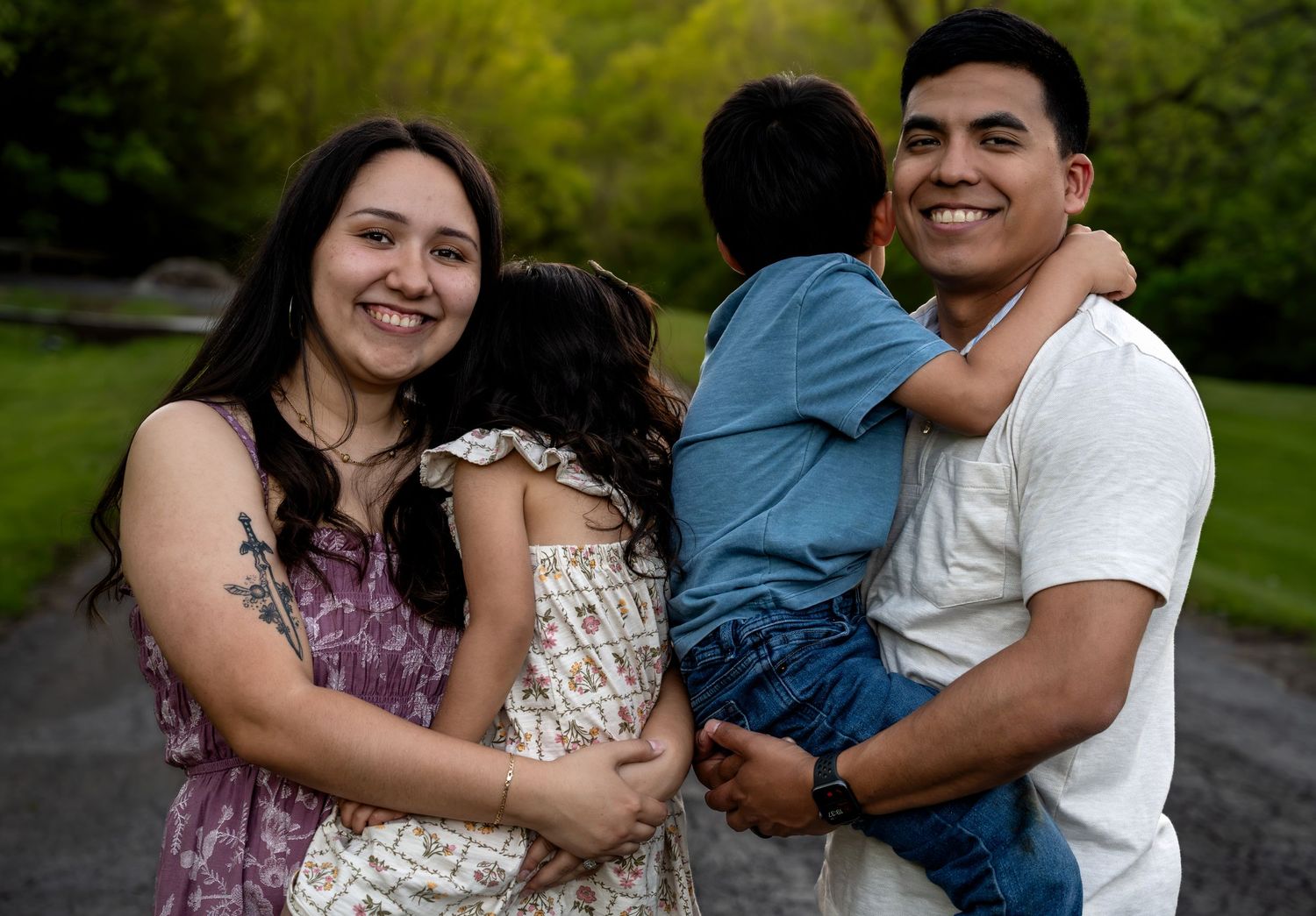 Happy family of four posing outdoors, parents smiling while holding their young children in a lush green park setting.