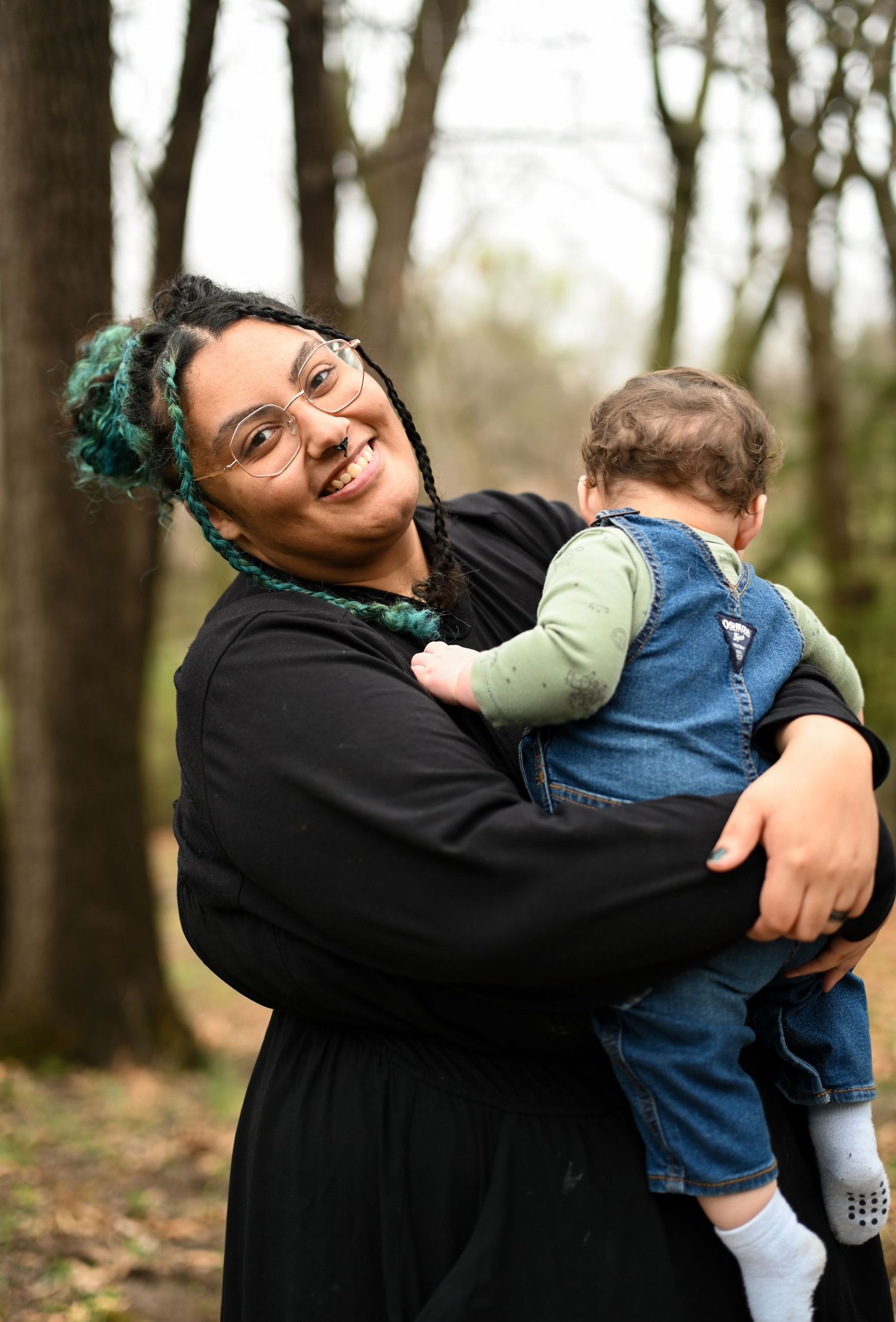 Happy mother with teal braided hair holding baby in denim overalls smiling in outdoor wooded setting.