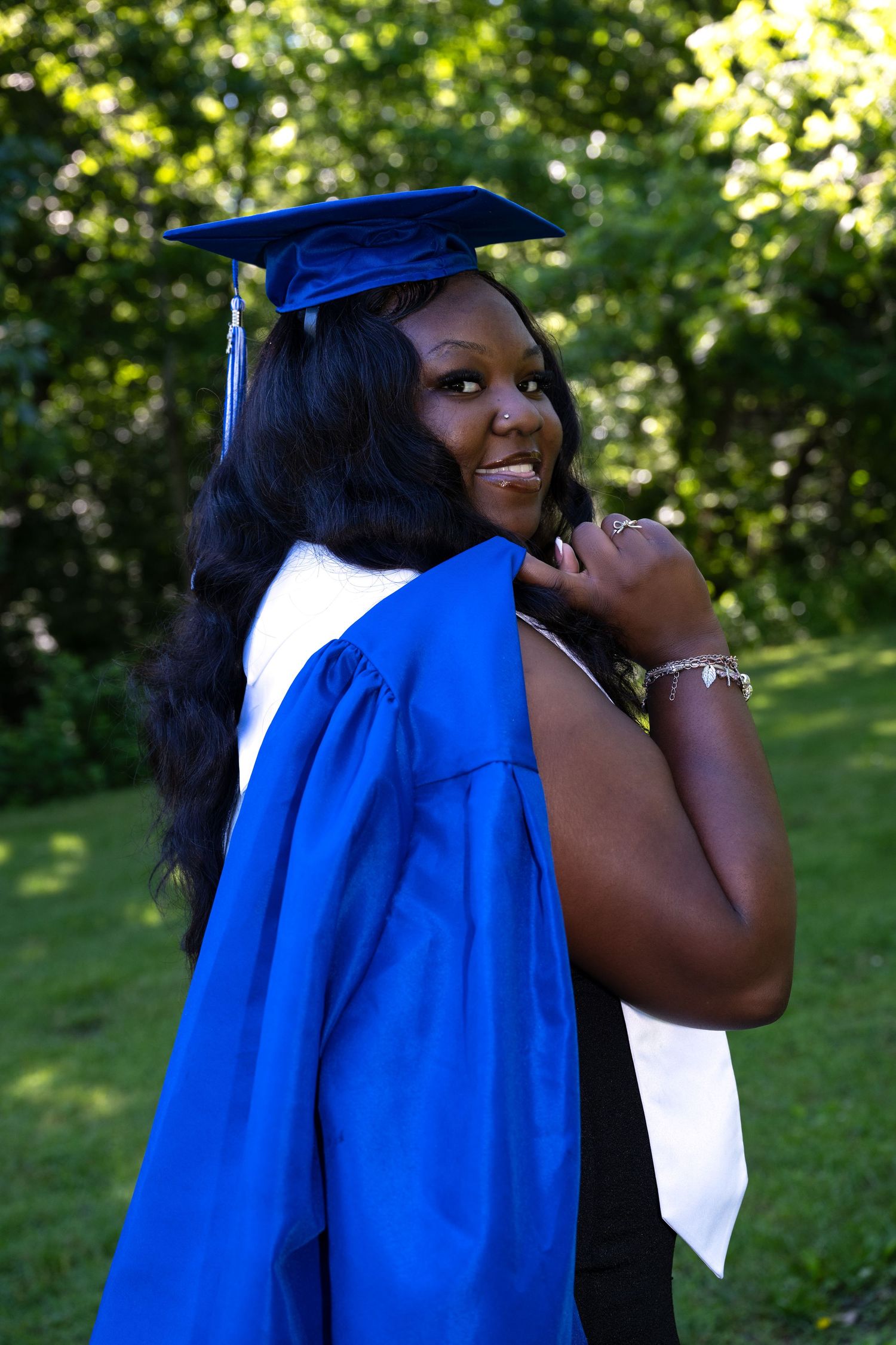portrait of a Smiling graduate in blue cap and gown glancing back over shoulder in a lush green outdoor setting.