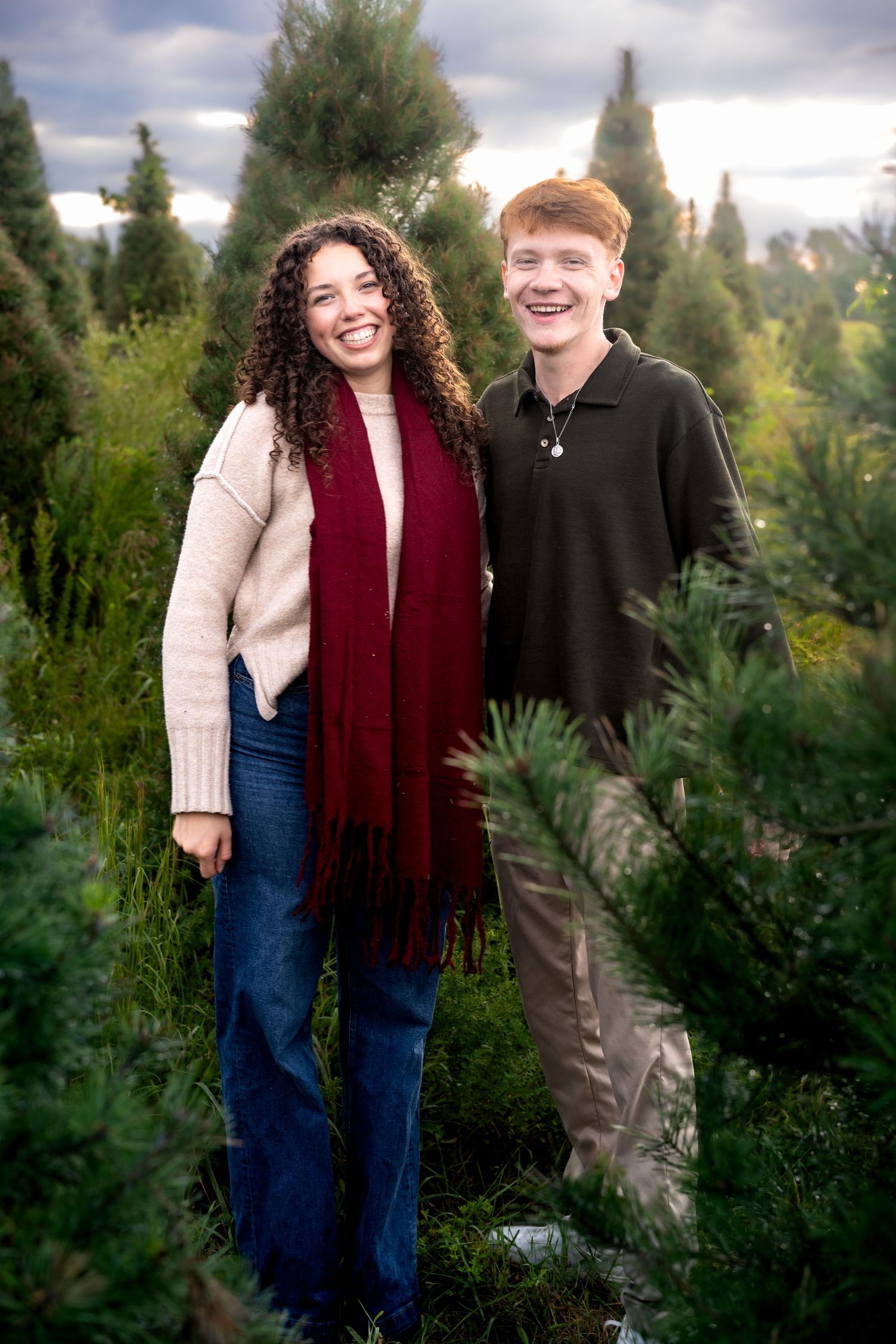A smiling young couple poses among green Christmas trees at a tree farm, woman in cream sweater and red scarf.