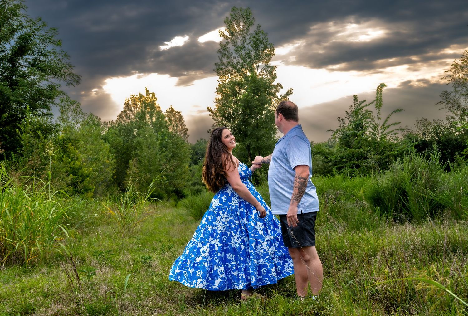 lifestyle portrait of a Couple dancing in a lush green field under dramatic stormy skies, woman in blue floral dress holding hands with man.