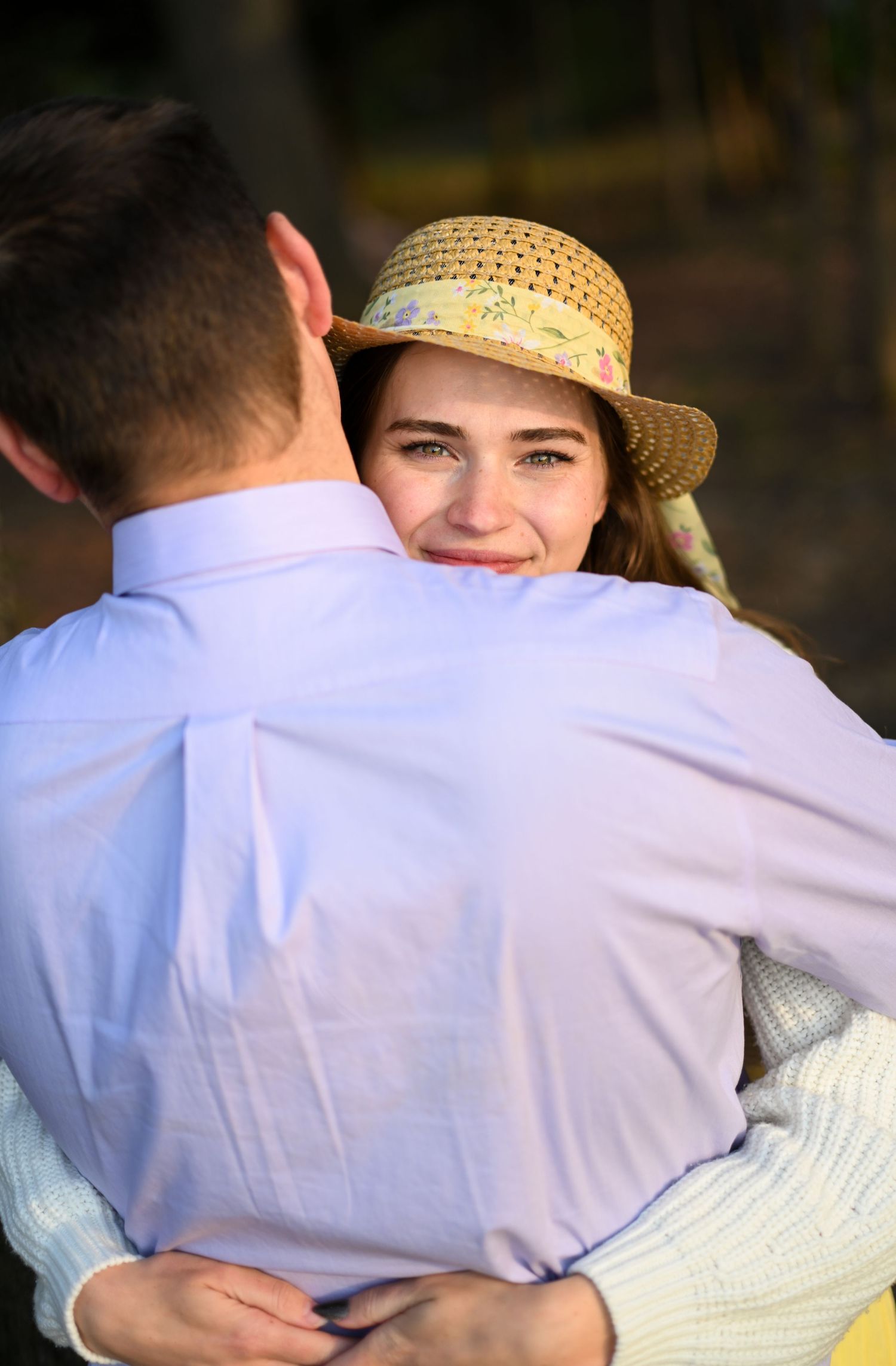 Smiling young woman in a straw hat hugging a man in a light blue shirt outdoors.