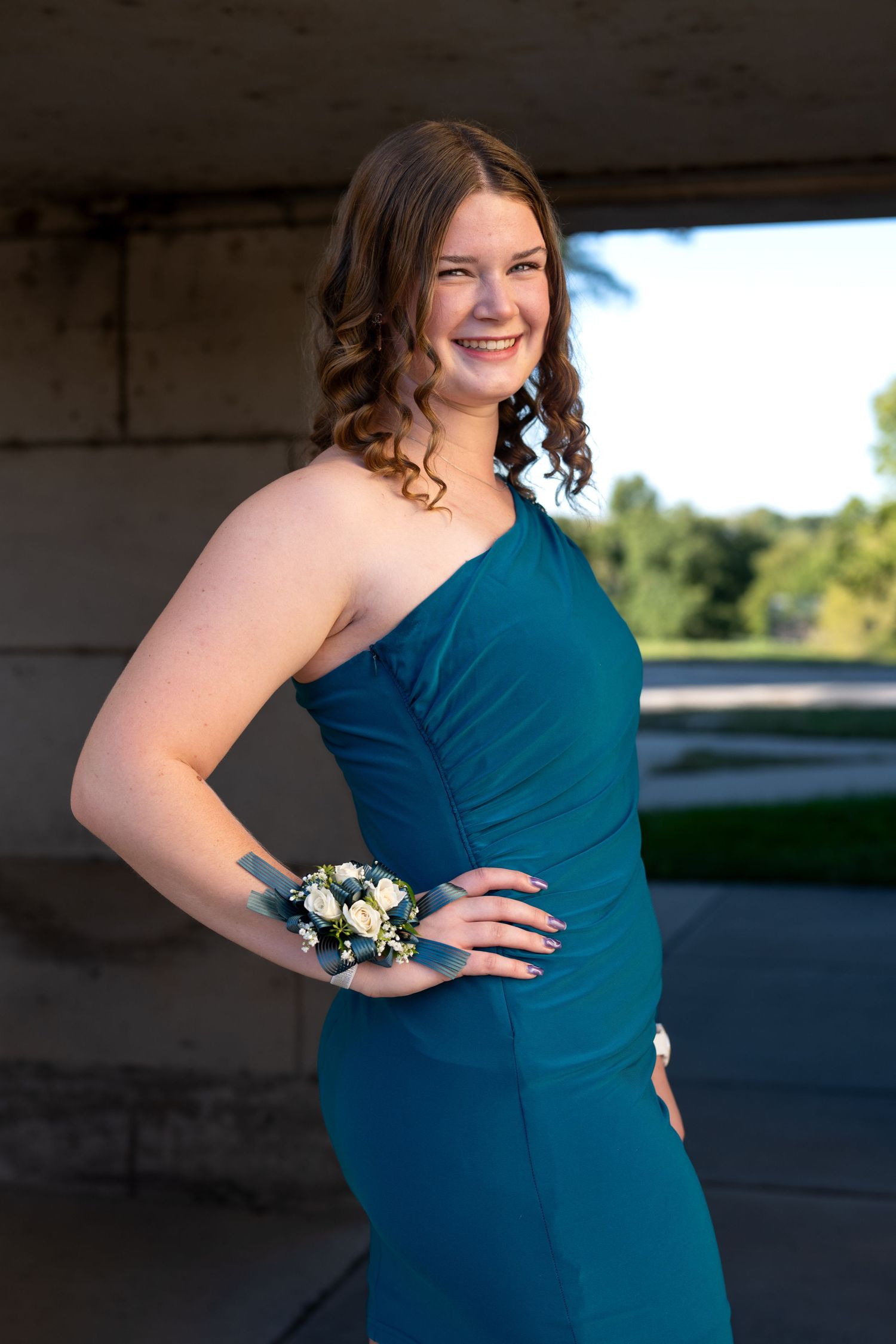 Young woman in teal one-shoulder gown with wrist corsage smiling at prom photo session outdoors.