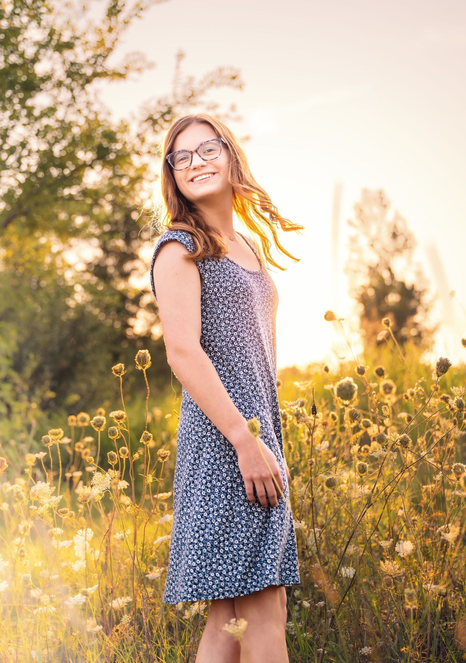 lifestyle portrait of a Smiling young woman in blue floral dress standing in sunny wildflower field at golden hour.