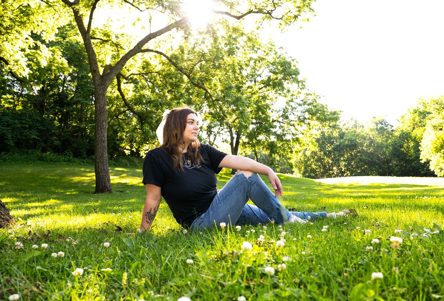 portrait of a Young woman in black shirt and jeans sitting relaxed on green grass under a sunlit tree outdoors.