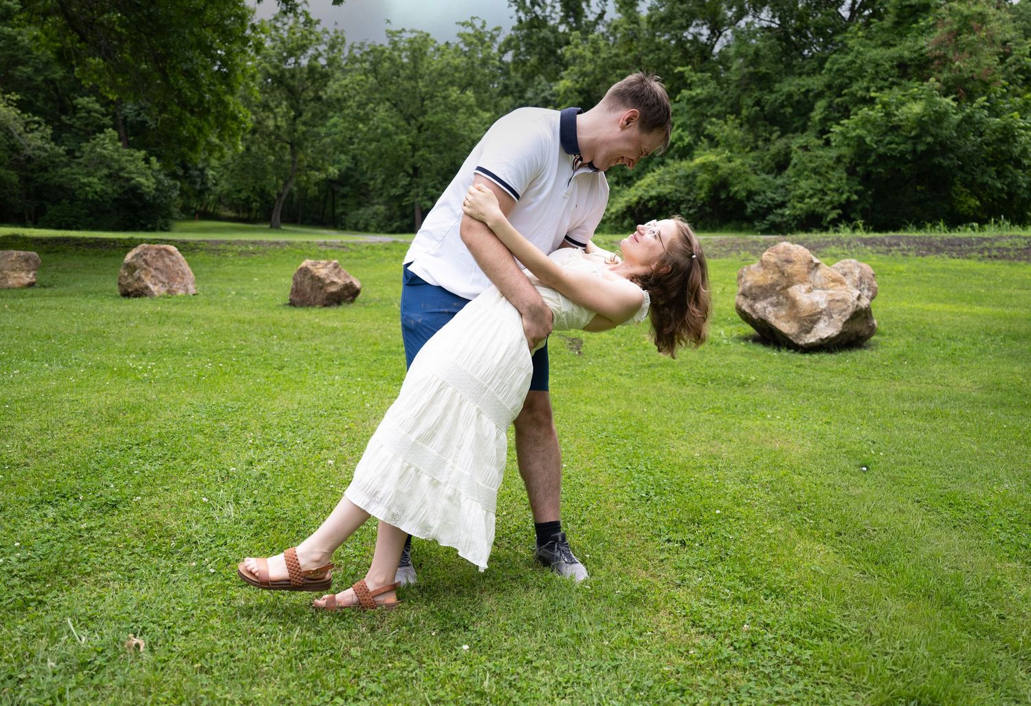 portrait of a Couple shares a romantic dip kiss on a lush green lawn surrounded by trees.