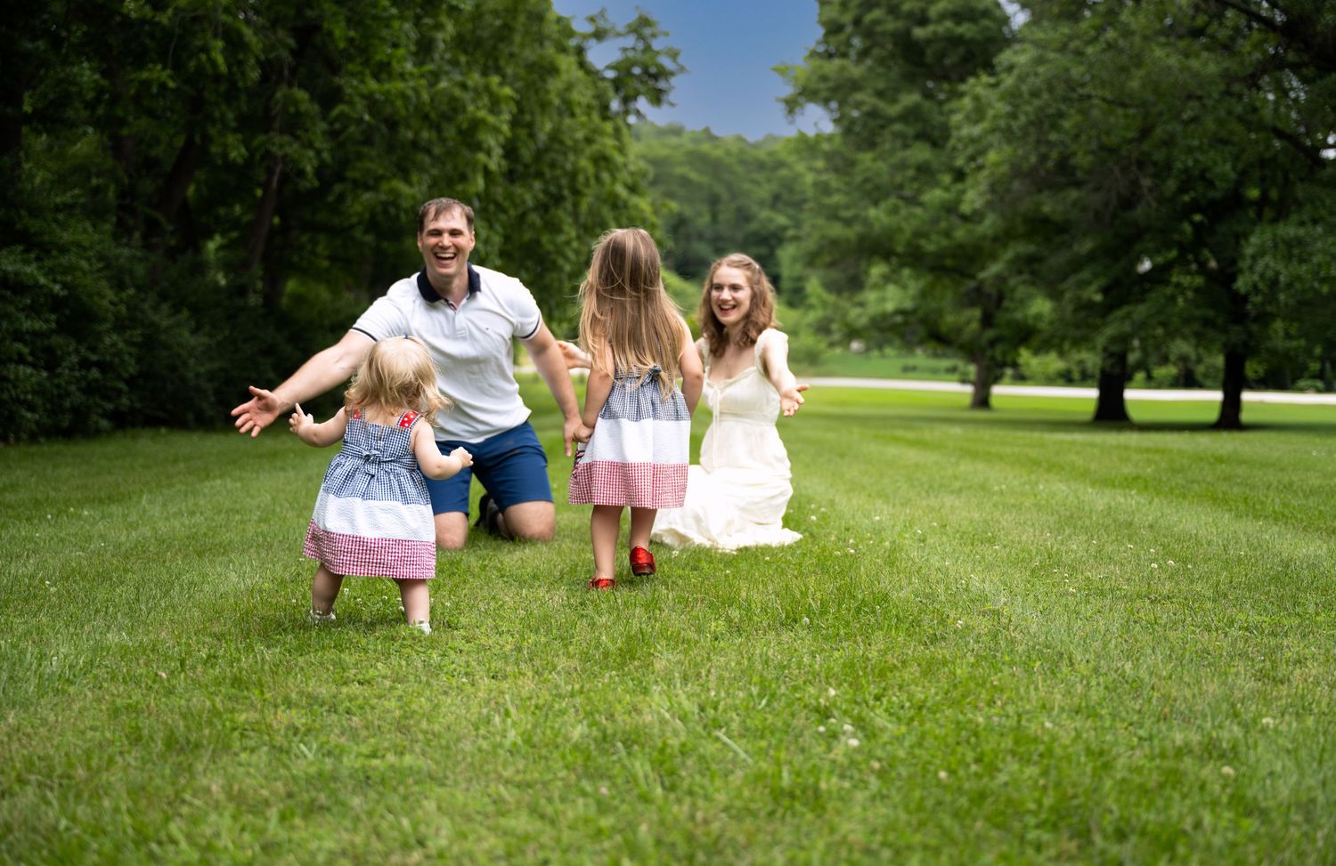 portrait of a Joyful family with two young girls in dresses running toward father on a lush green lawn.