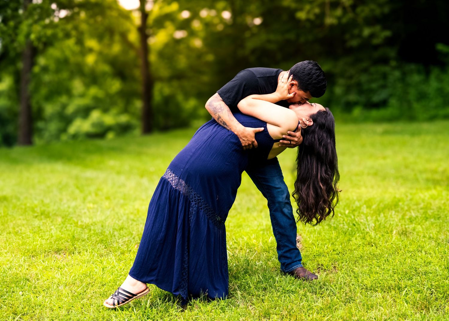 lifestyle portrait of a Couple sharing a romantic dip kiss on green grass in a park, woman in navy blue dress.