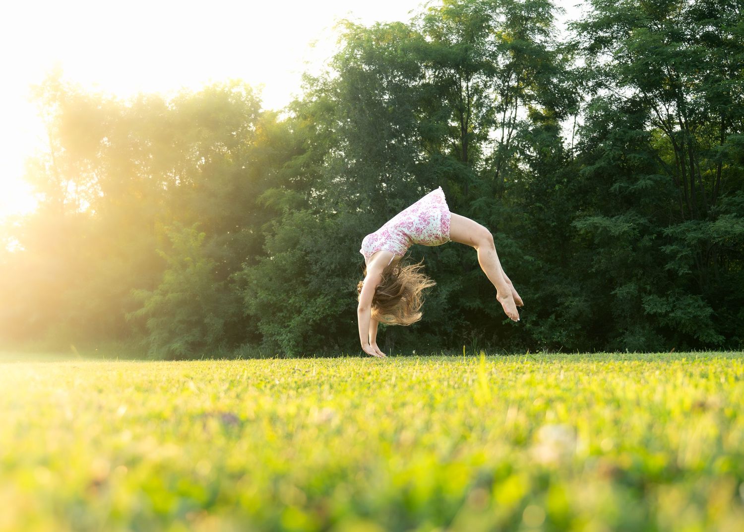 Young girl doing a backflip on lush green grass in a sunlit park surrounded by trees.