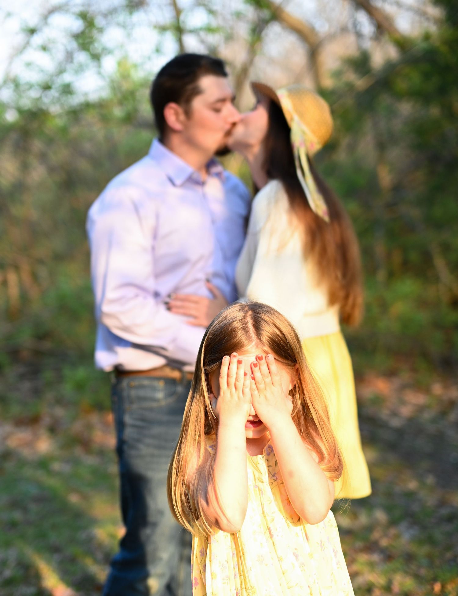 Young girl covering eyes as parents share a kiss in a sunlit outdoor background.