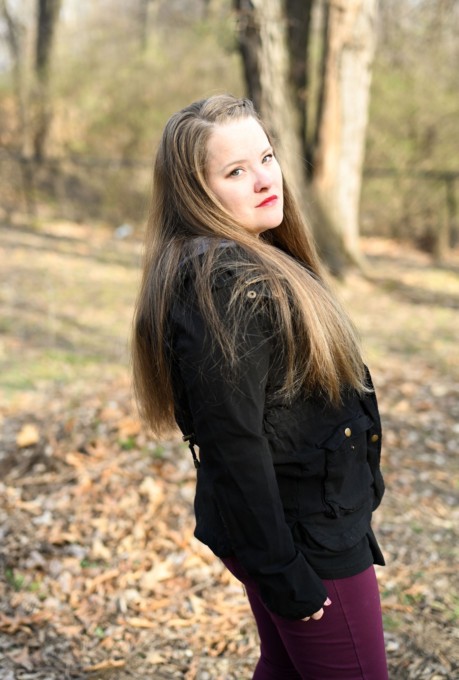 Young woman with long hair in black jacket glancing back over shoulder in woods.