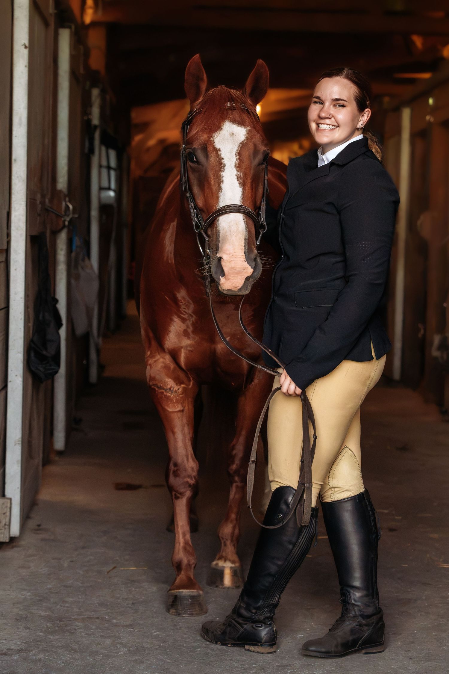 Smiling equestrian in black jacket and tall boots stands beside a chestnut horse in a stable aisle.