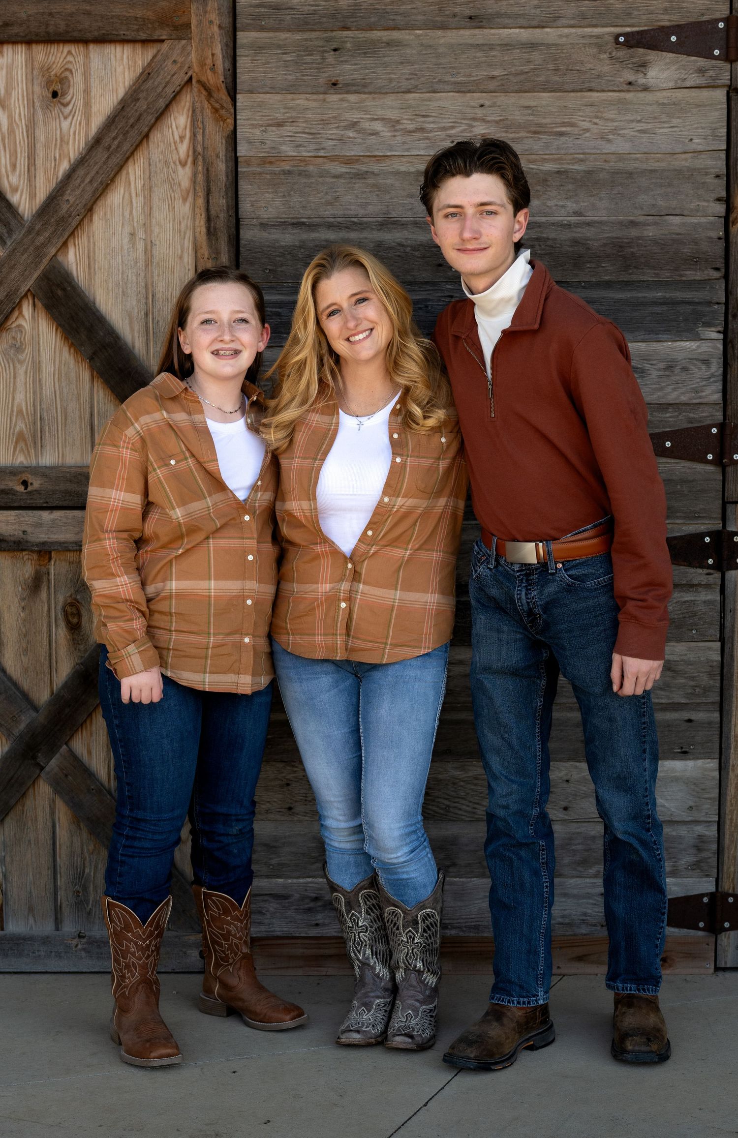 Three young people in matching rust plaid shirts, jeans and cowboy boots pose arm in arm in front of a rustic wooden barn door.