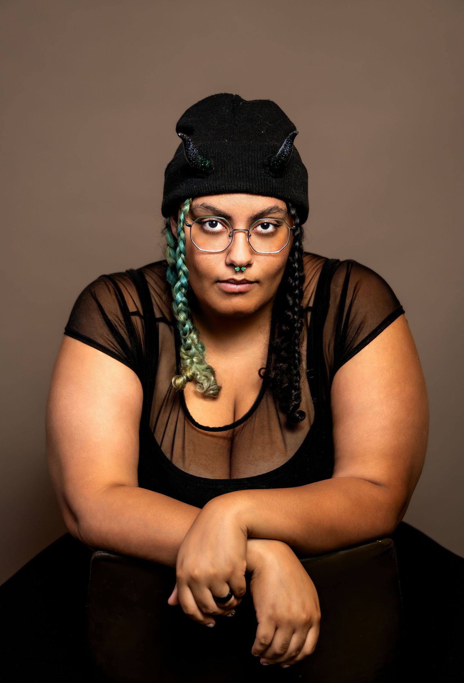 Person with green braids, black beanie, glasses, and sheer top posing confidently in a studio portrait.