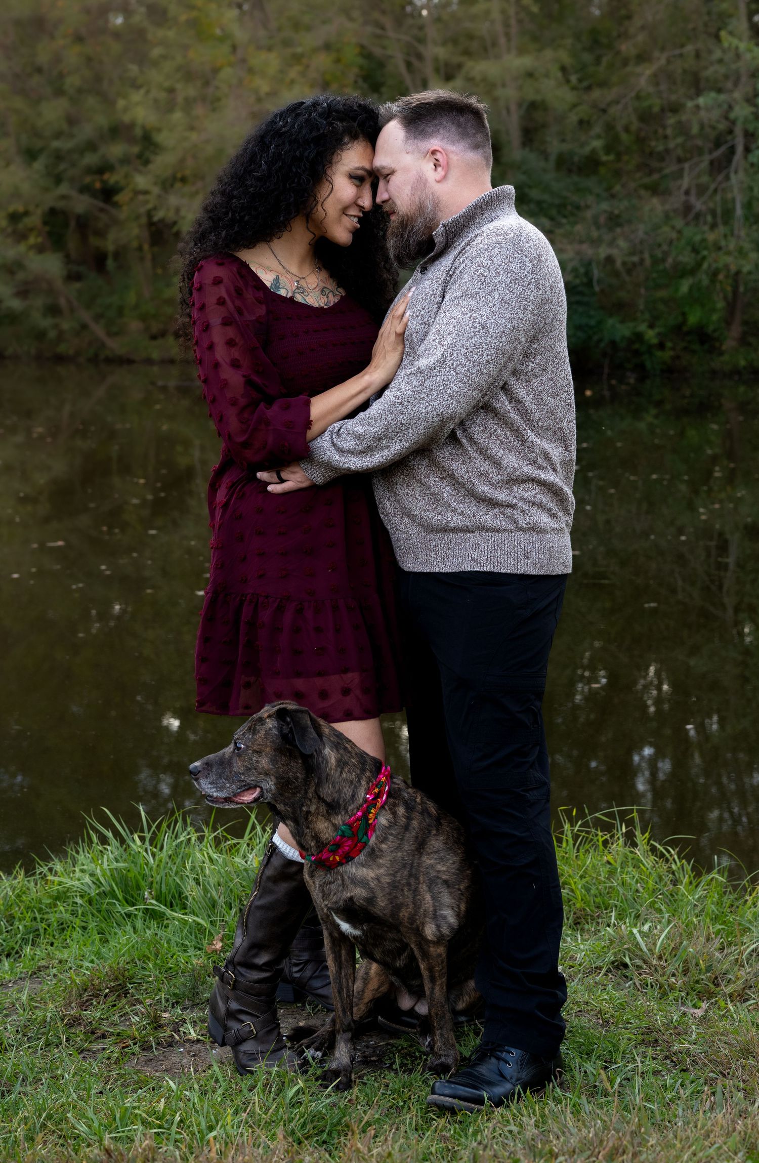 A couple embraces affectionately beside a calm pond while a brindle dog wearing a red collar sits at their feet on the grass.