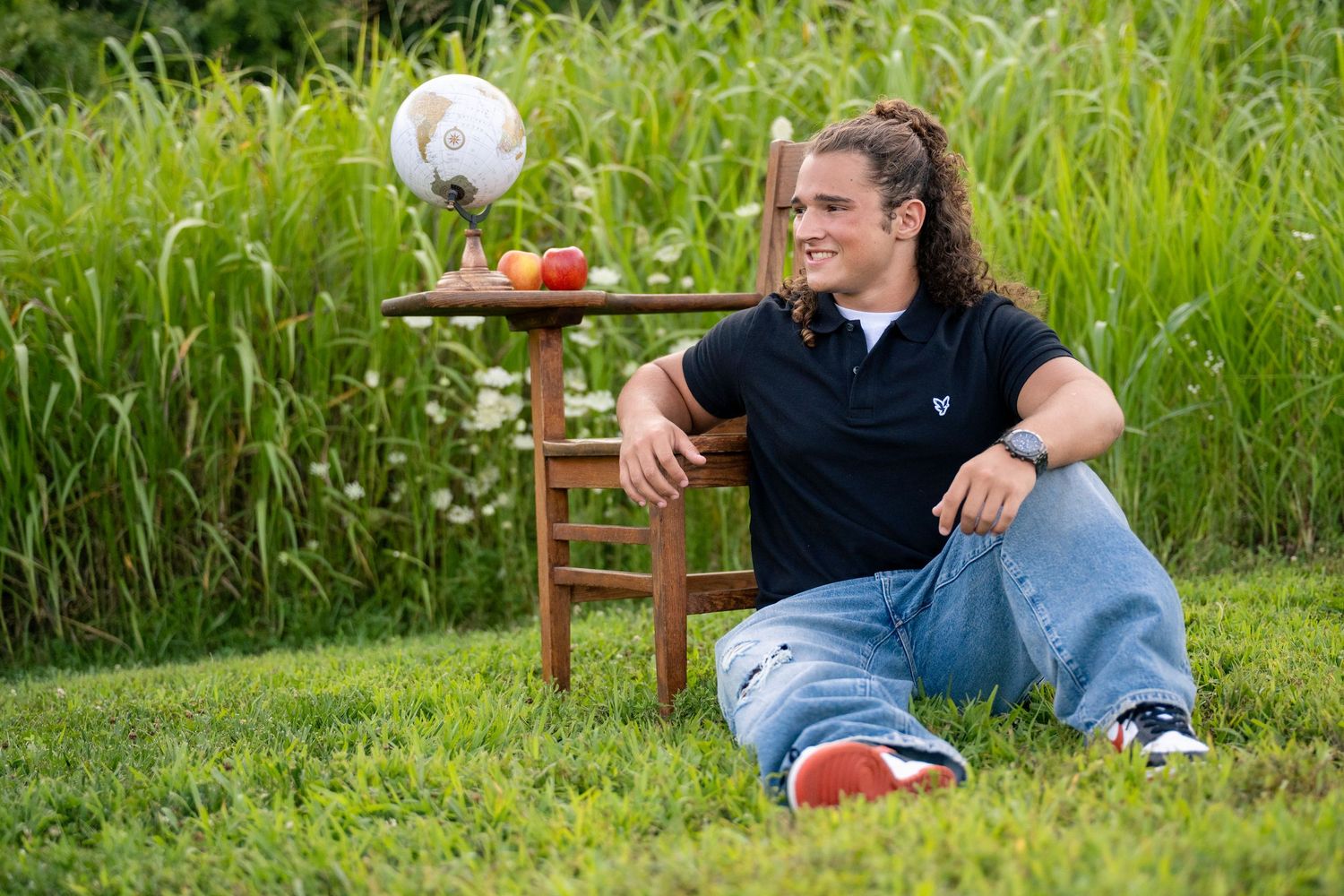 portrait of a young man with curly hair sits on grass smiling near school desk  outdoors.
