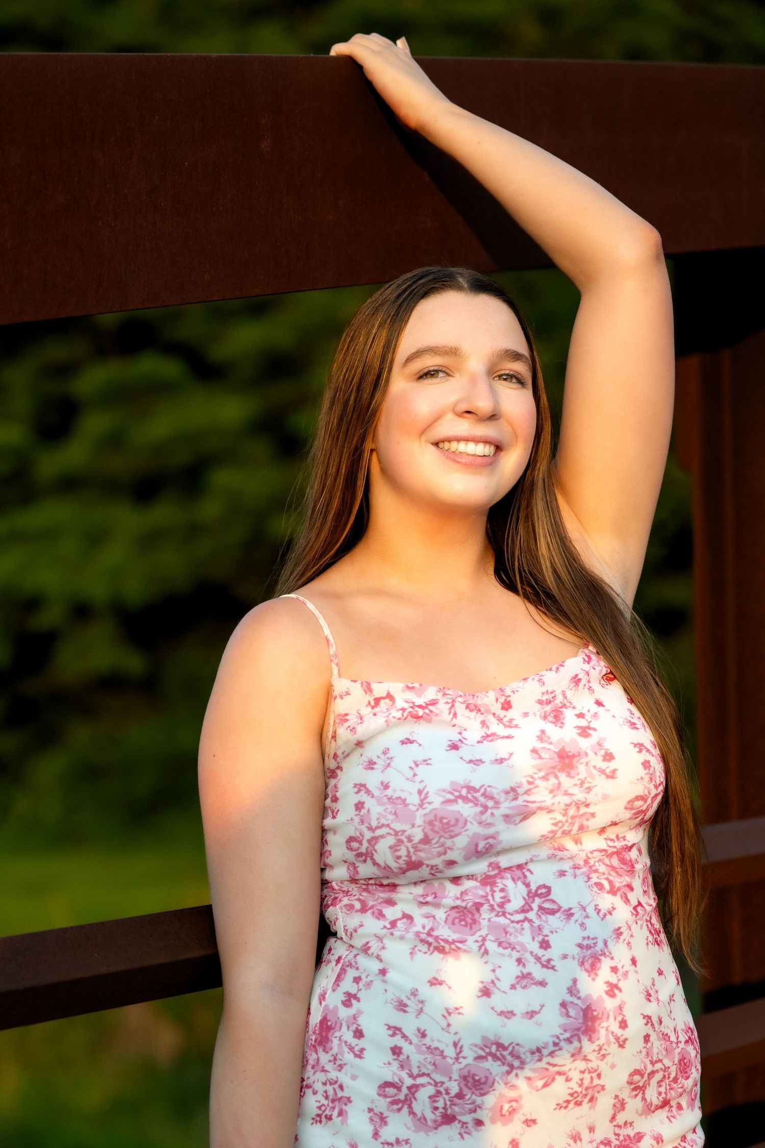 Young woman in floral sundress smiling outdoors, long brown hair, leaning against wooden fence in golden hour light.