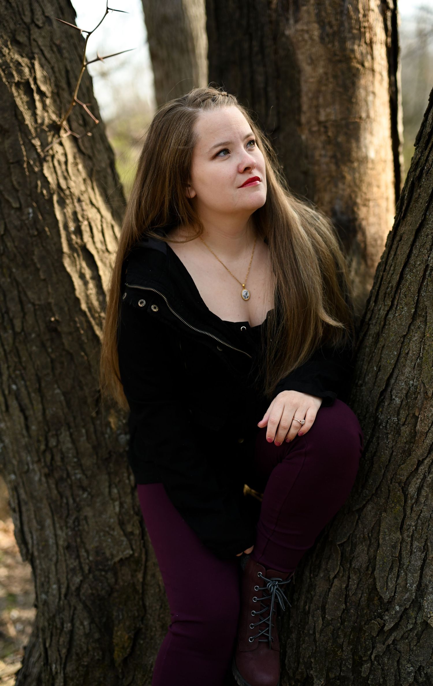 Young woman with long brown hair in black jacket and burgundy pants posing between two trees in a forest.