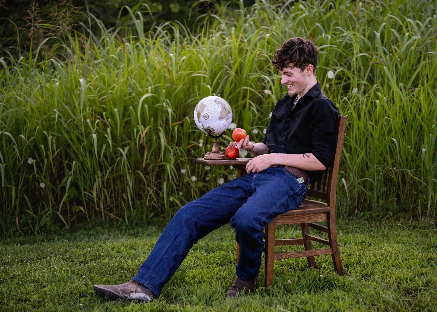 portrait of a Young man seated in wooden chair outdoors examining an apple  with globe on desk