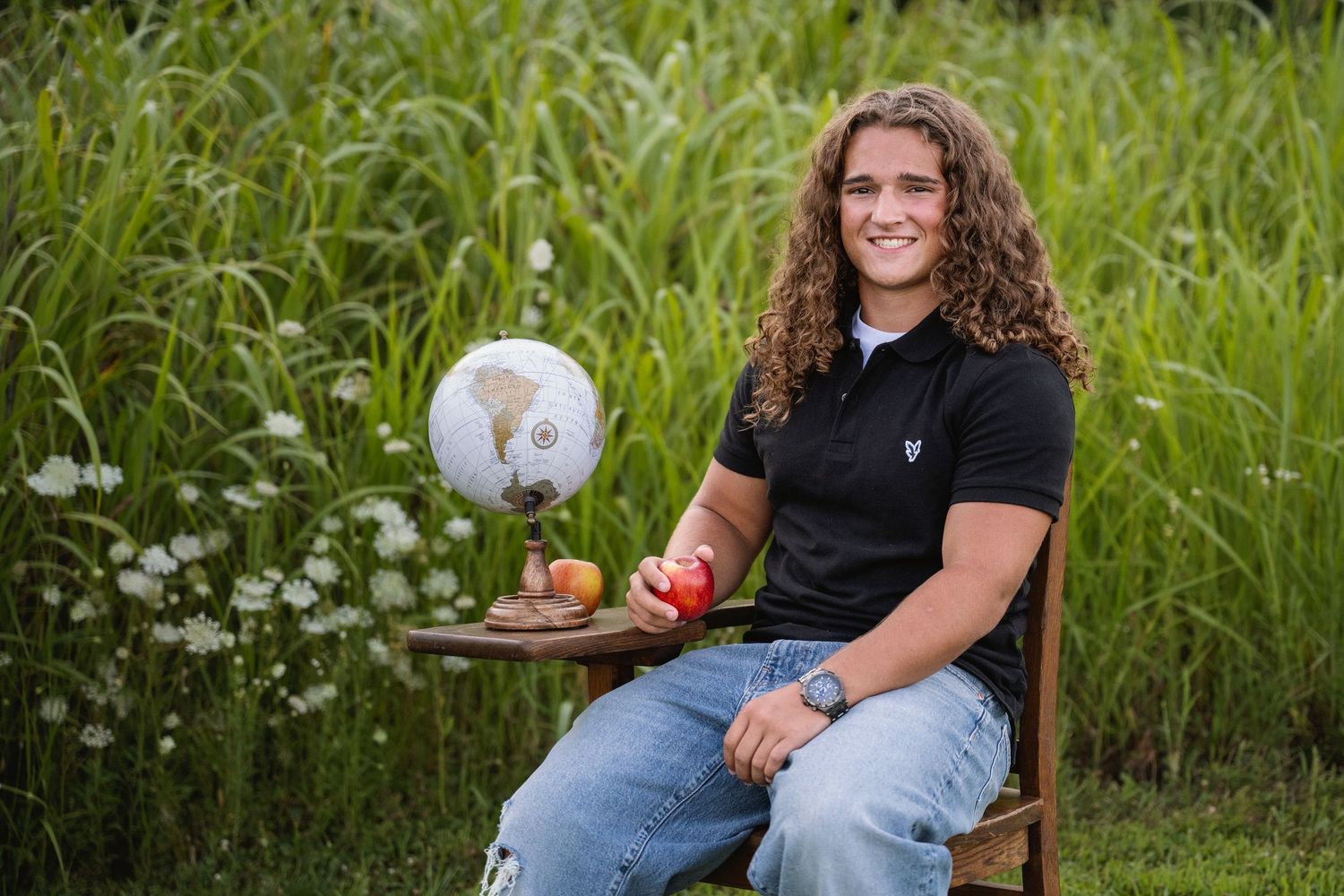 portrait of a young man with curly hair smiles holding red apple beside globe in nature.