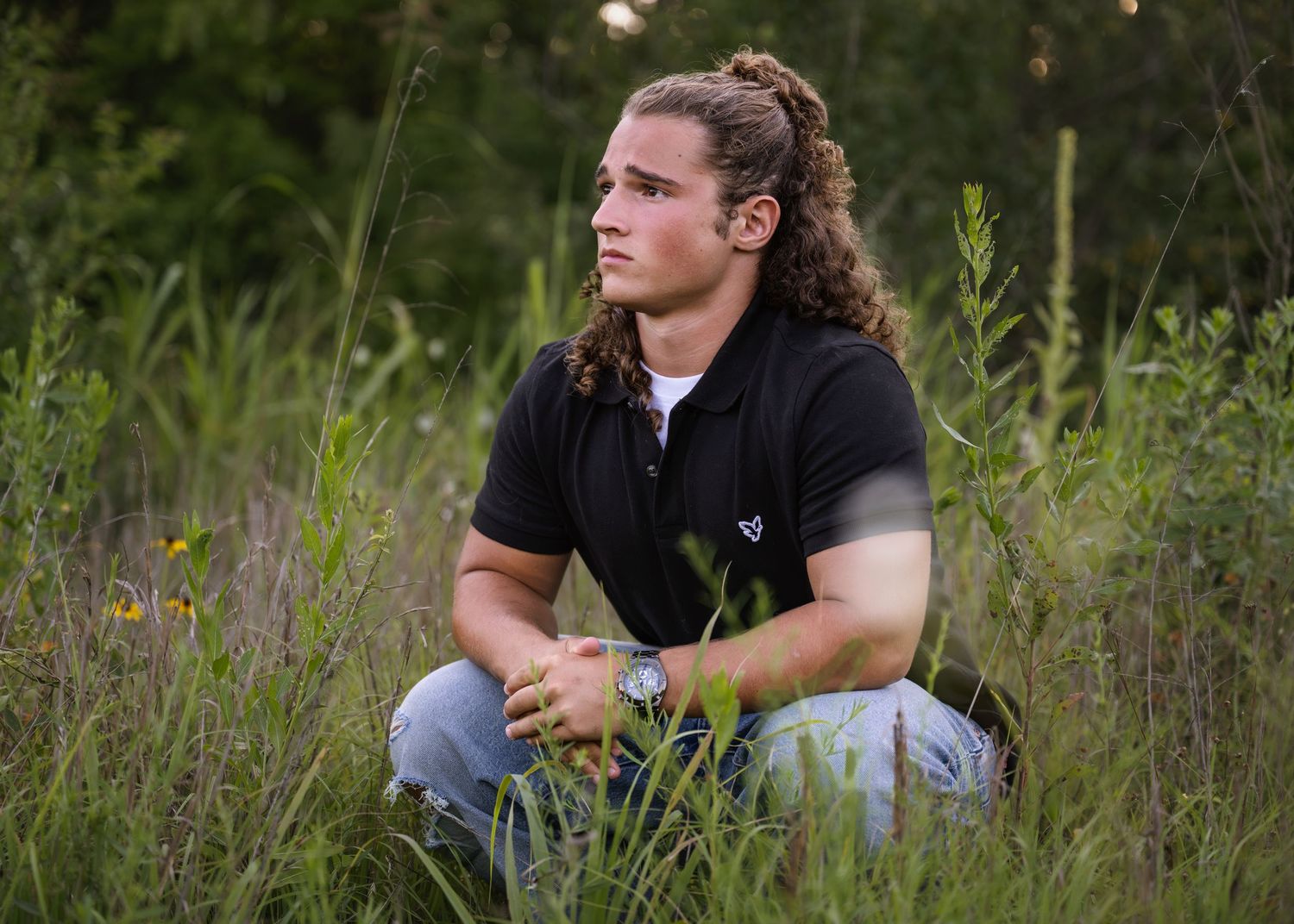 portrait of a young man with pulled-back curly hair sits thoughtfully in tall green grass.
