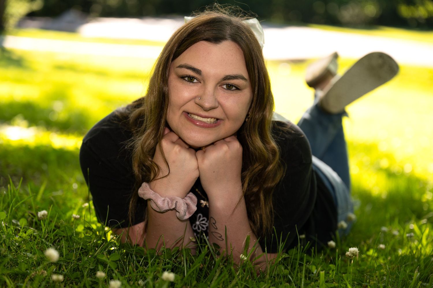 Young woman with brown hair lying on green grass smiling with hands under chin on a sunny day.