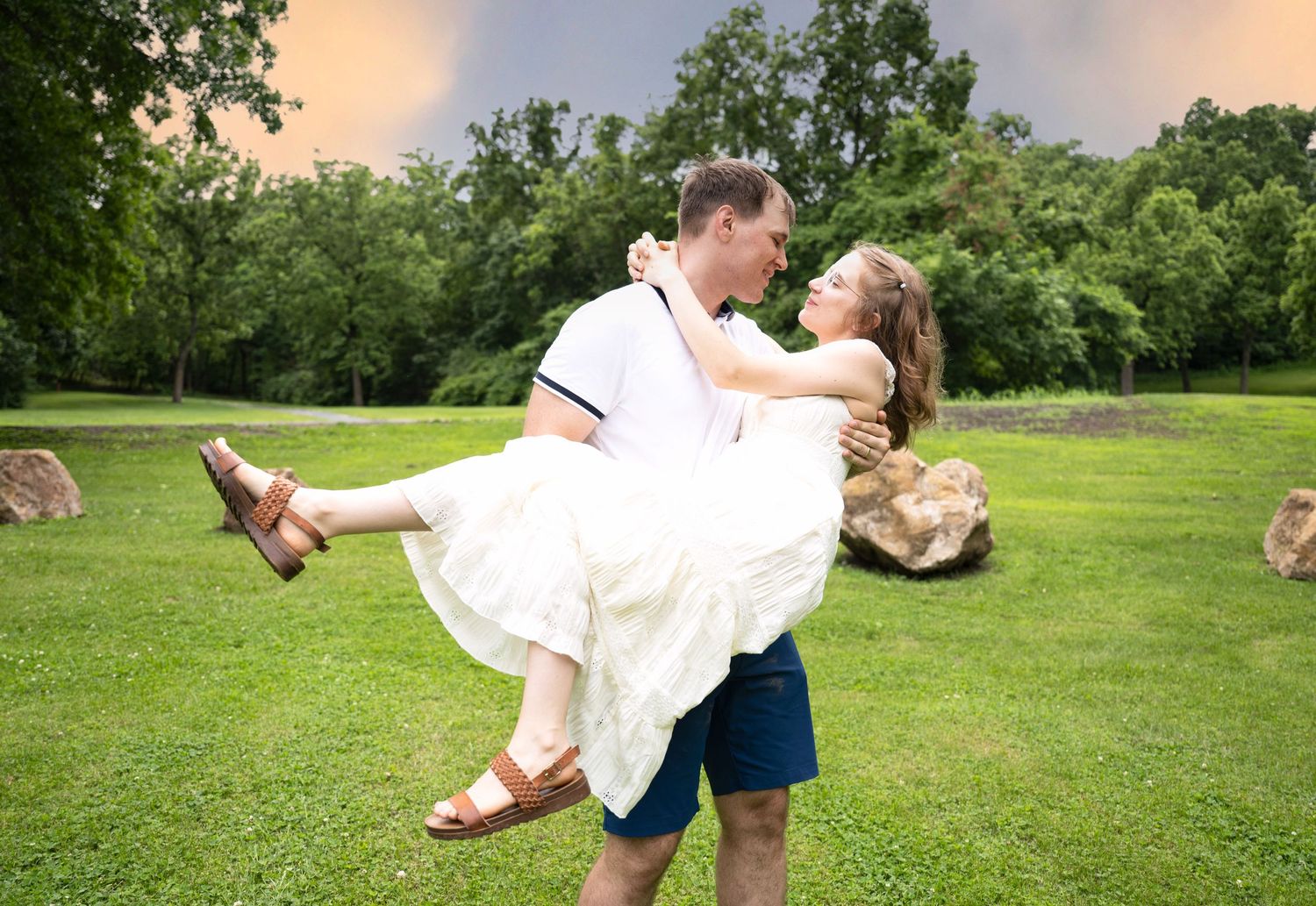portrait of a Man lifts woman in white dress for a kiss in a scenic park with dramatic sunset sky.