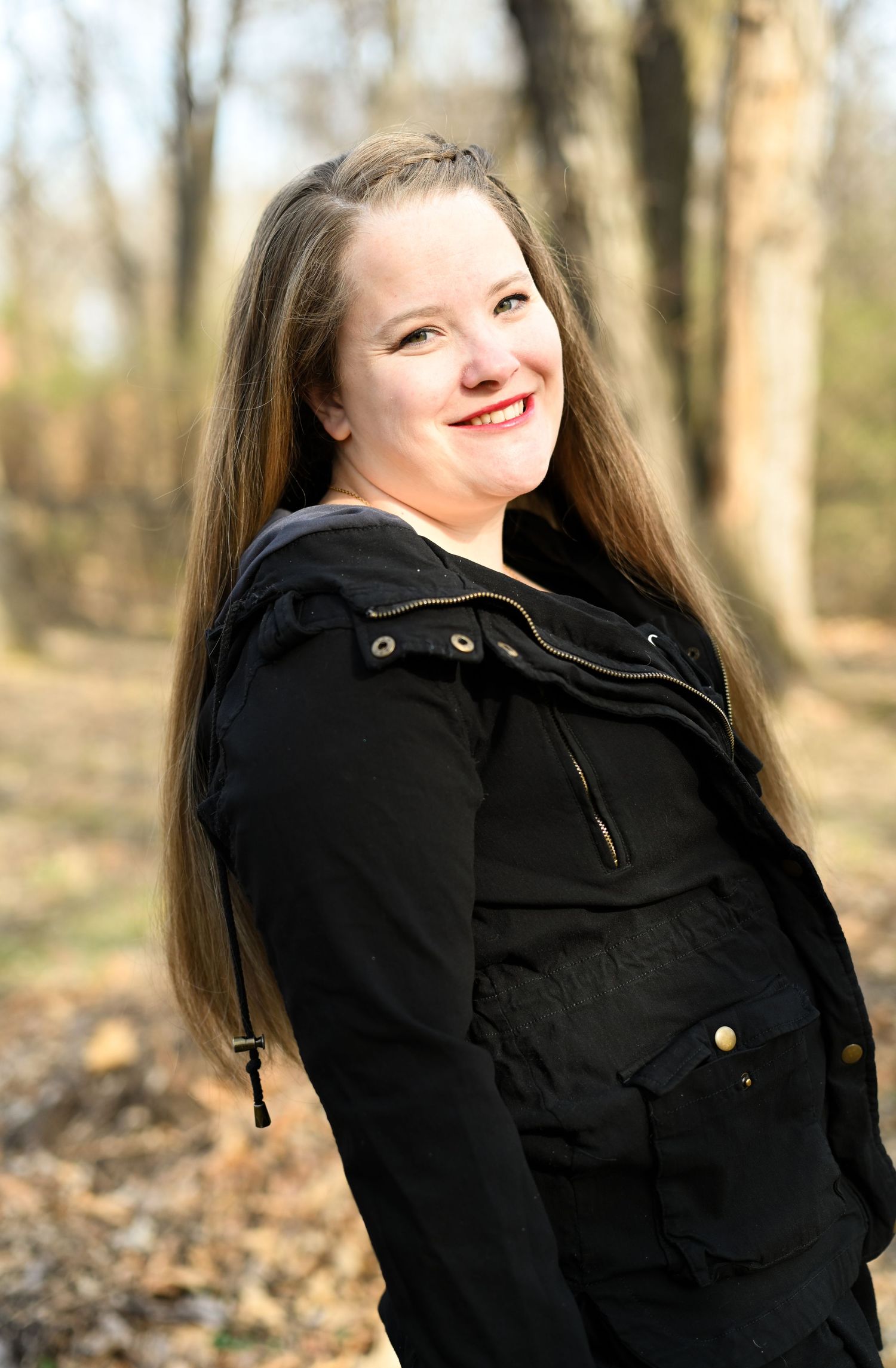 Cheerful young woman with long hair in black jacket smiling in autumn forest.