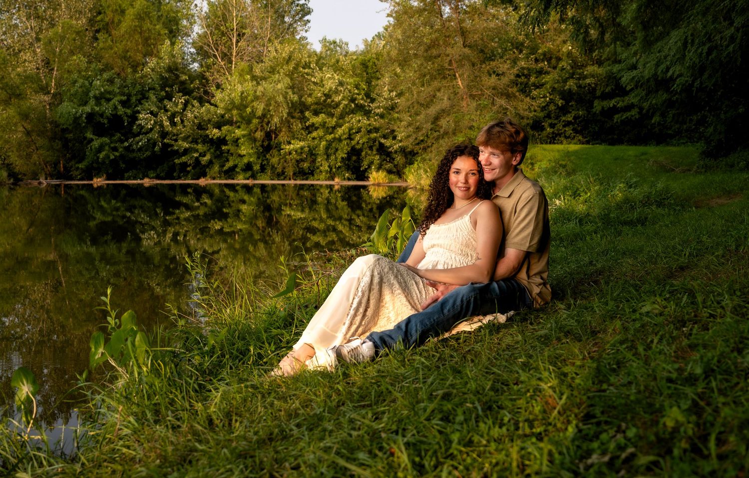lifestyle portrait of a Couple sitting together by a tranquil pond surrounded by lush green trees in warm golden sunlight.