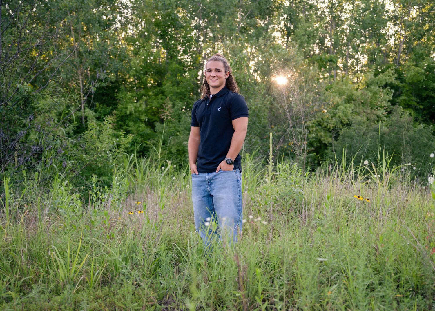 portrait of a young man with long hair stands in sunlit meadow wearing black polo shirt.
