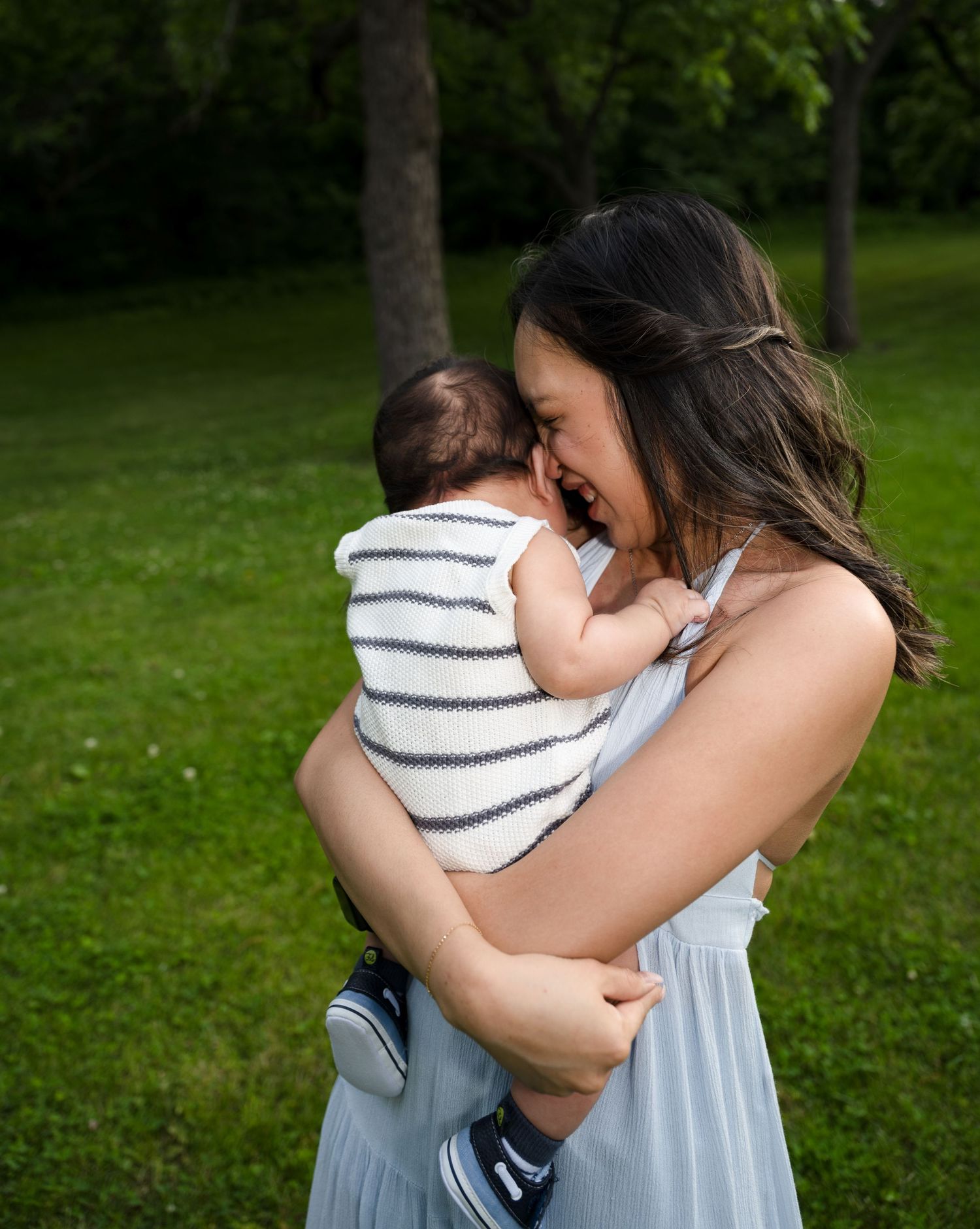 portrait of a Joyful mother in light blue dress laughing and nuzzling her infant baby outdoors in a green park.