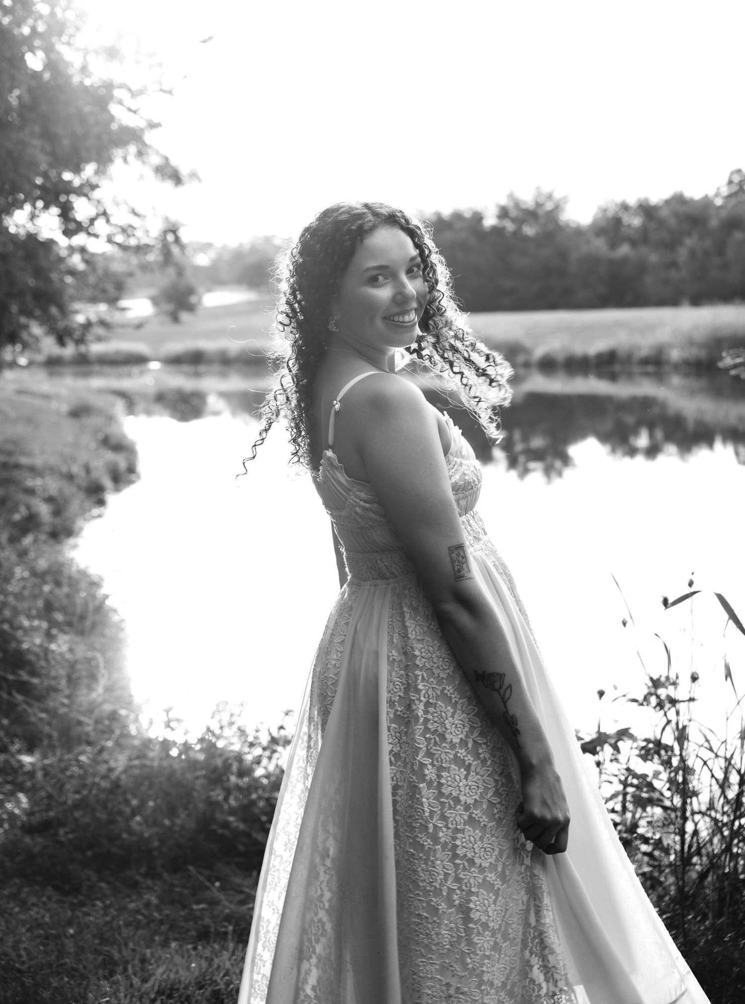 Black and white lifestyle portrait of a smiling woman in a lace gown posing by a serene pond surrounded by trees.